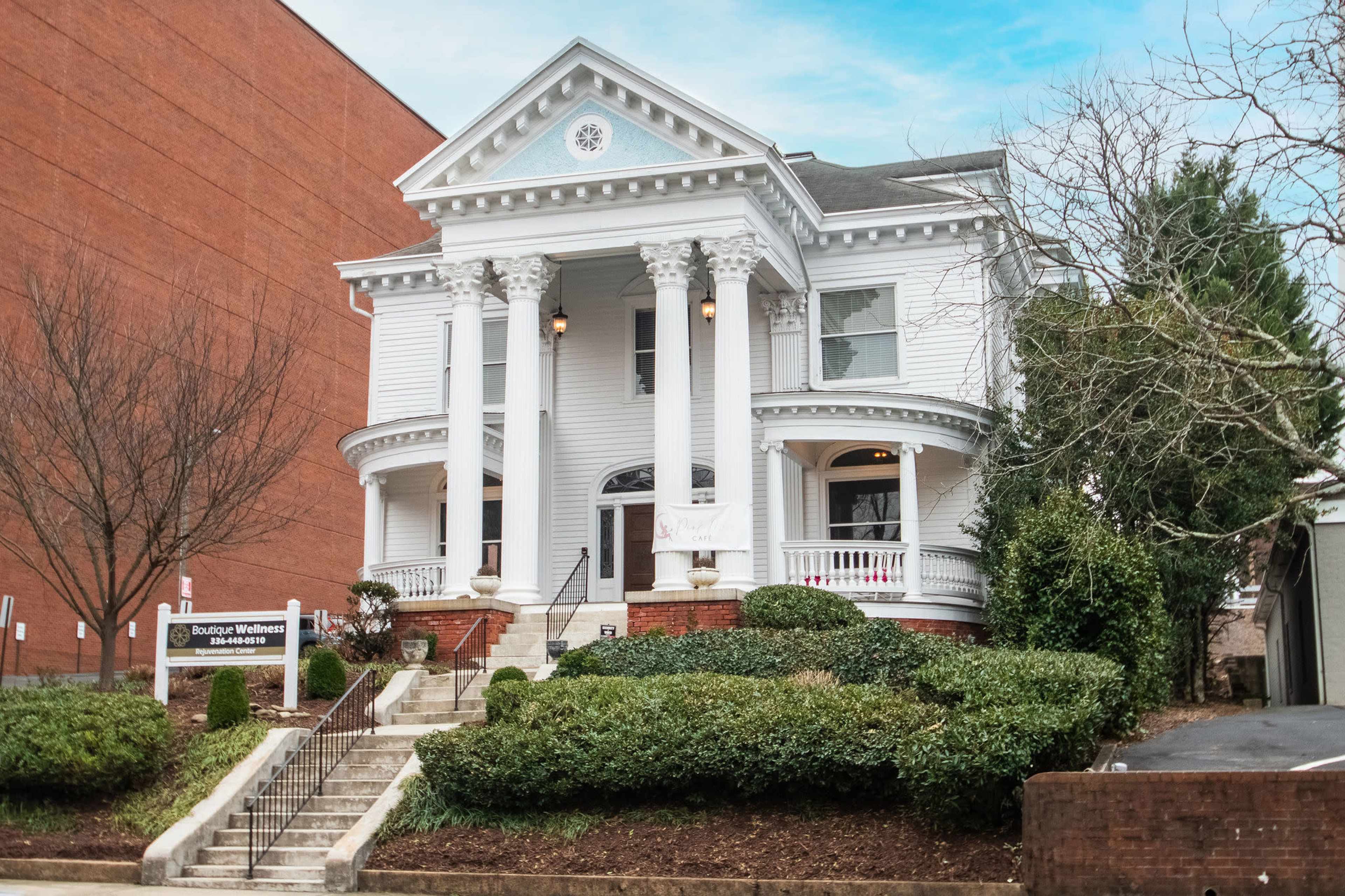 A historic white mansion with columns and a staircase, surrounded by neatly trimmed shrubs and trees.