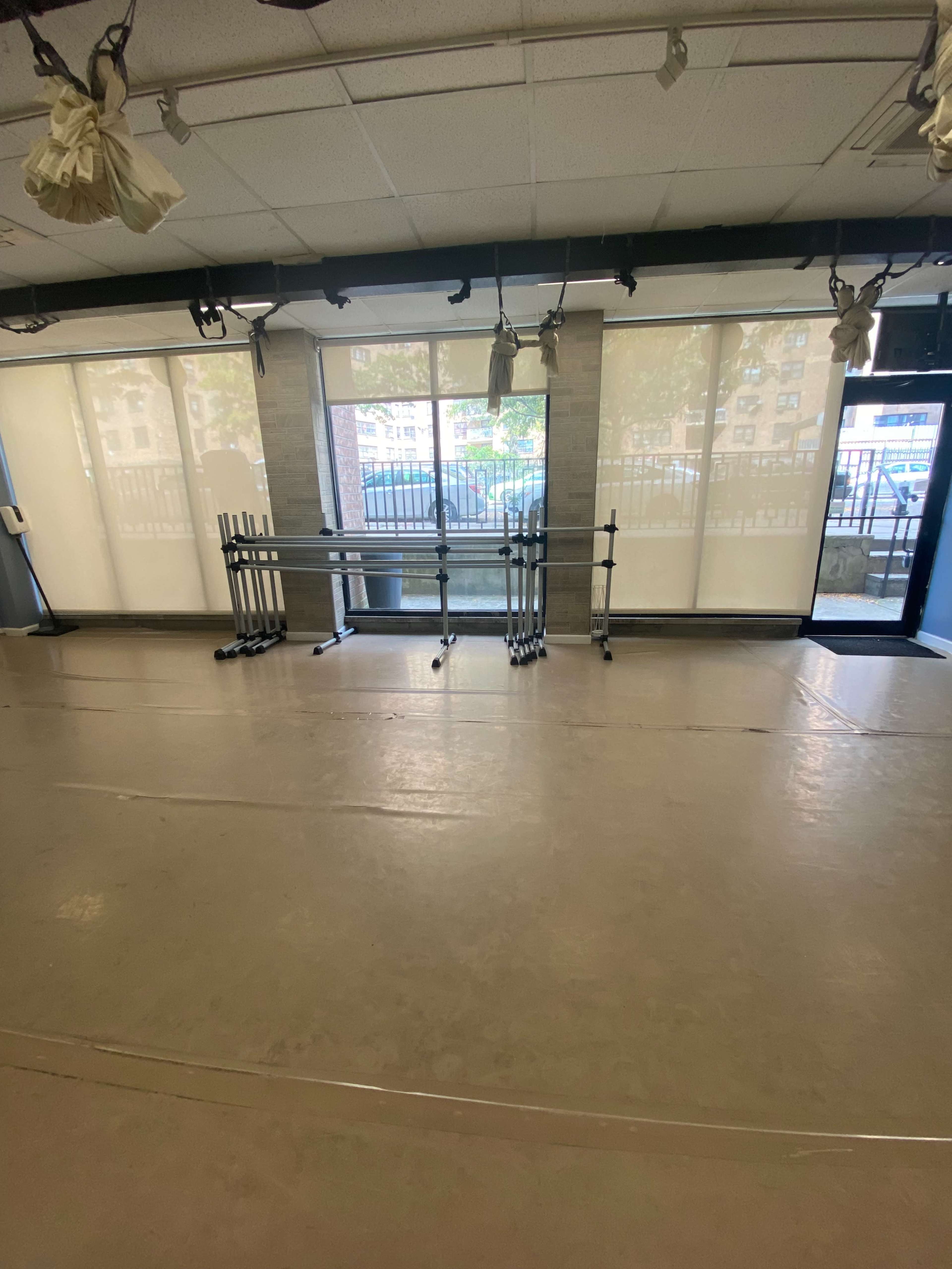 The image shows a spacious room with wooden floors and large windows that allow natural light, featuring a row of ballet barres against a wall.