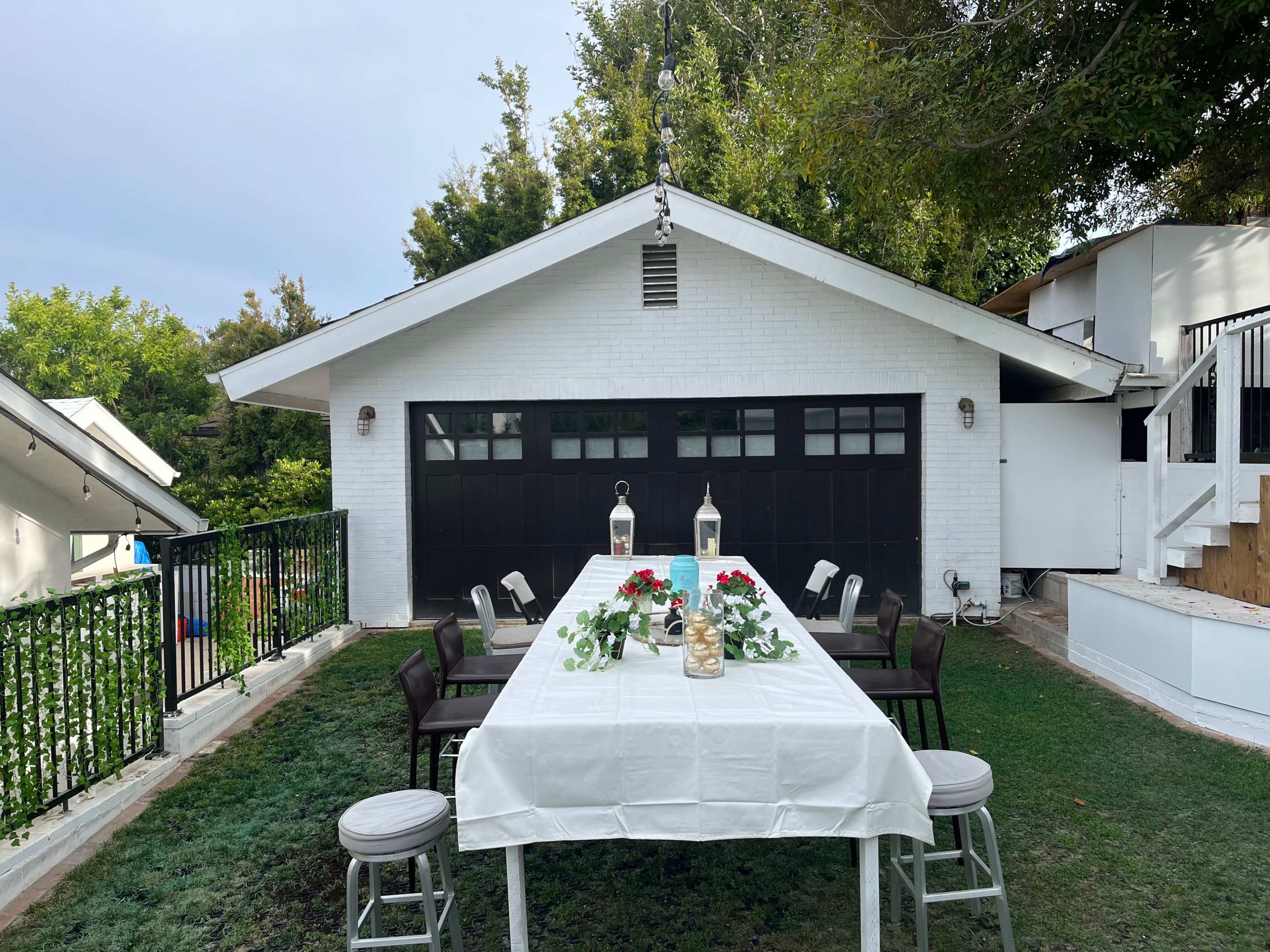 A long table is set outside in front of a white garage, surrounded by green grass and trees.