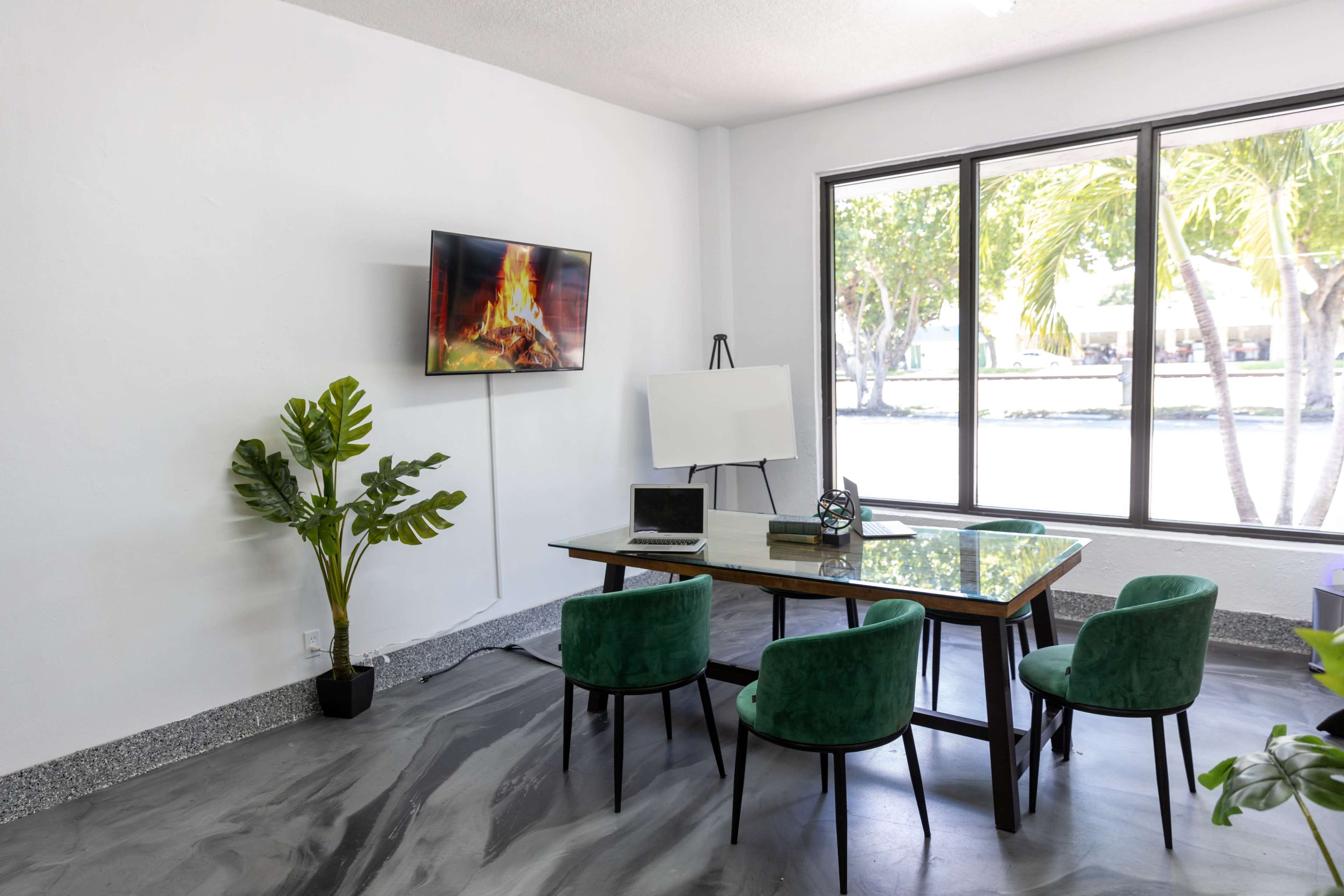 A modern conference room features a glass table surrounded by green chairs, a television mounted on the wall, and a whiteboard on an easel.