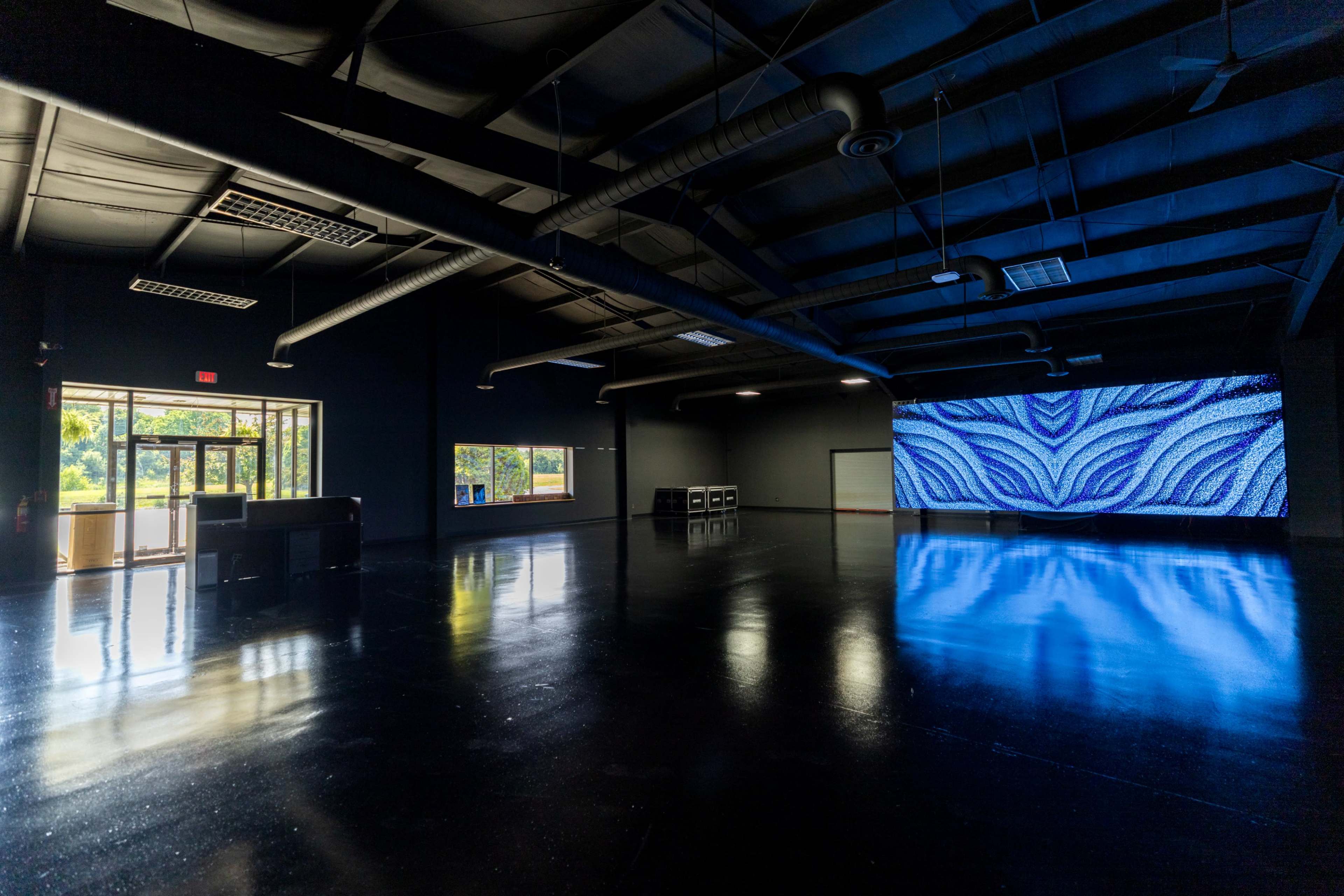 The image shows a spacious, empty room with glossy black floors, large windows letting in natural light, and a vibrant, illuminated screen on one wall.