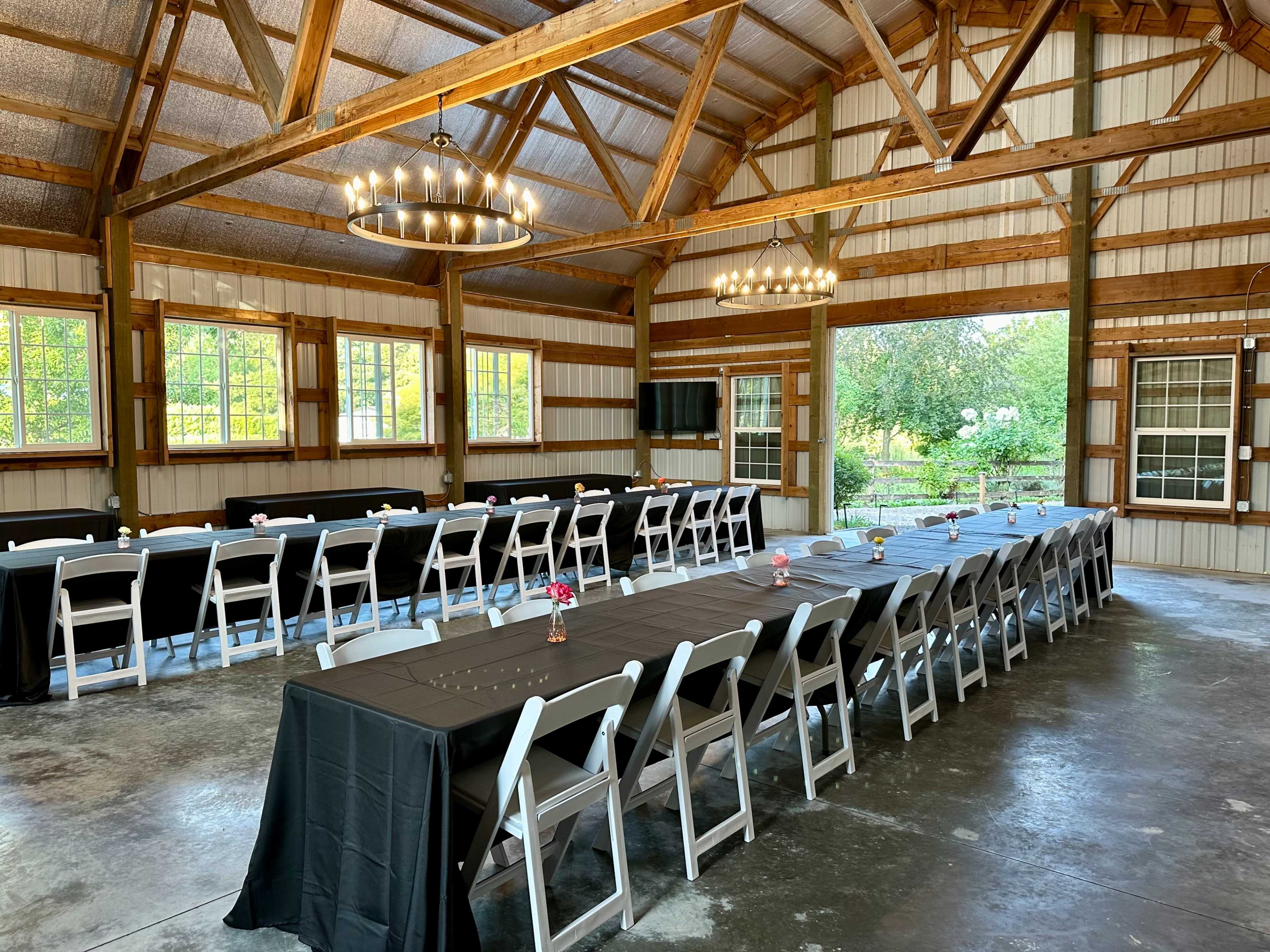 The image shows a spacious event room with wooden beams, arranged with long tables covered in black tablecloths and white chairs, set up for a gathering.