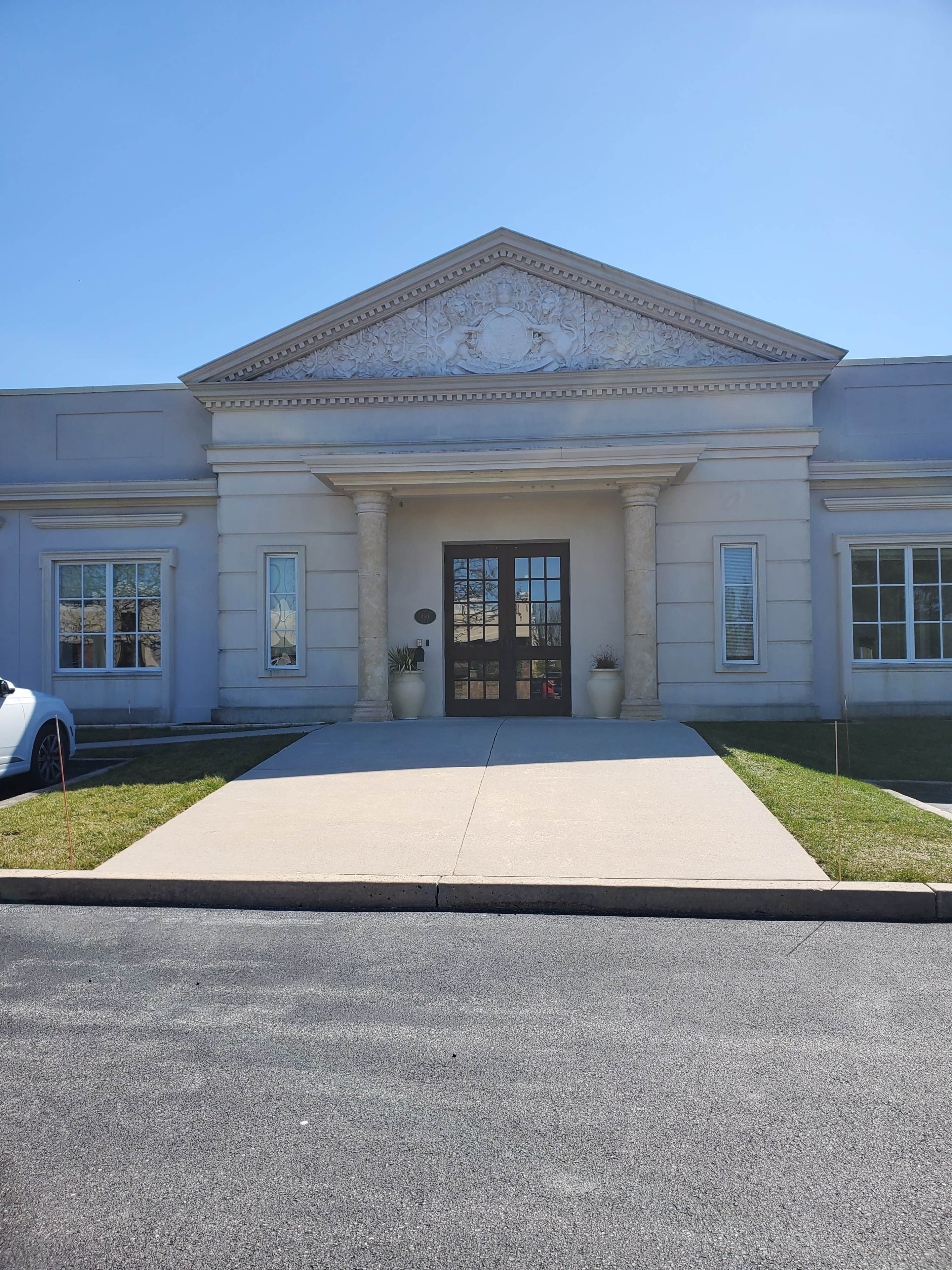 The image shows a classical-style building with a prominent entrance featuring decorative columns and a sculpted pediment, set against a clear blue sky.