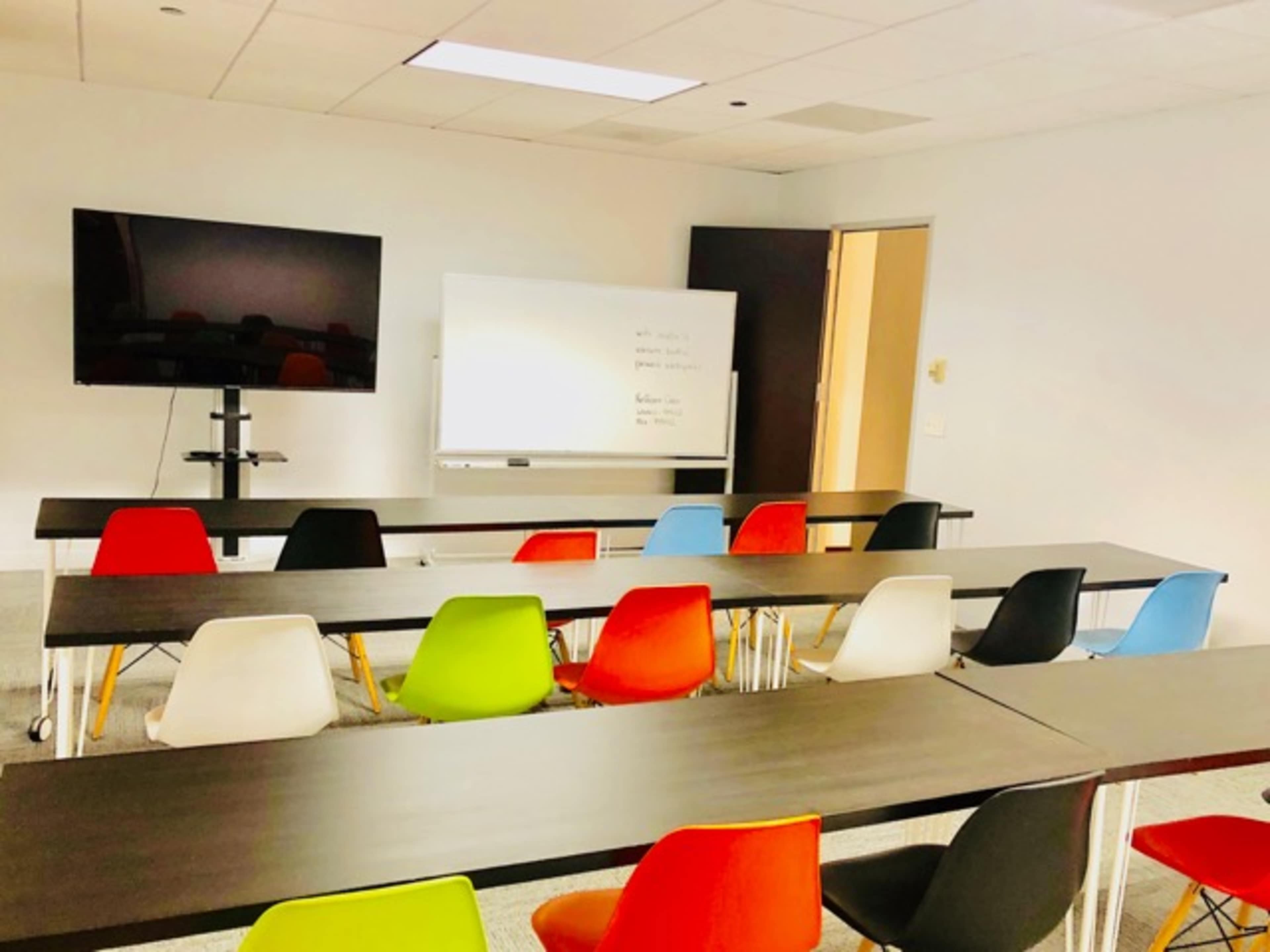 A brightly lit conference room features multiple tables arranged in rows, with colorful chairs and a large screen on one wall.