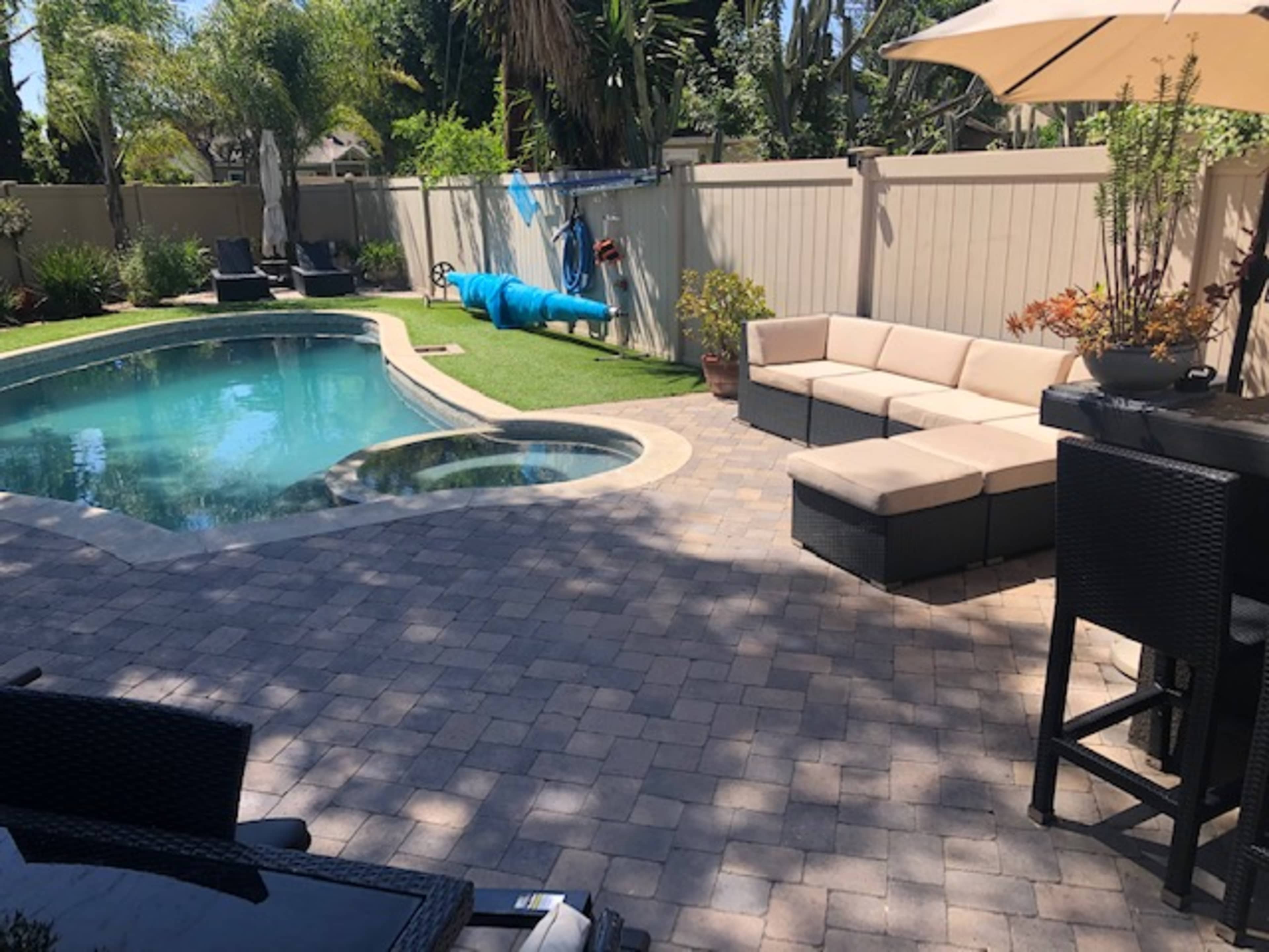 The image shows a landscaped backyard with a swimming pool, a spa, and a seating area featuring a sectional couch and tall bar stools under a patio umbrella.