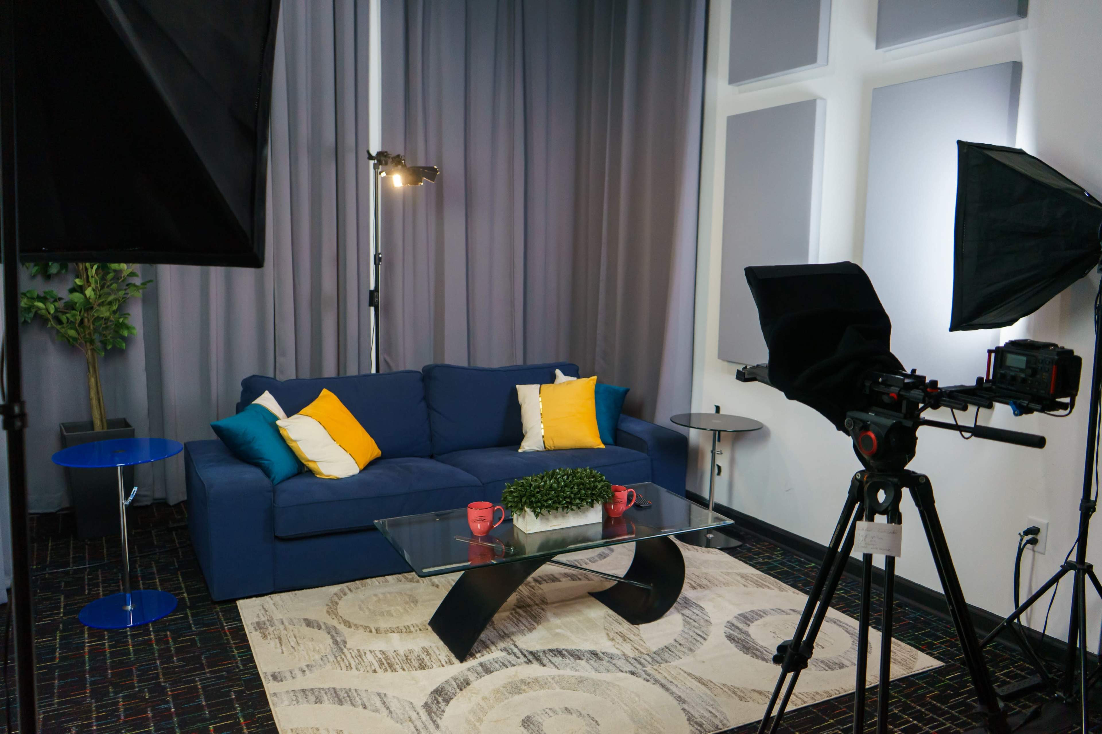 A modern living room setup with a blue sofa, decorative pillows, a glass coffee table, and a camera on a tripod facing the seating area.