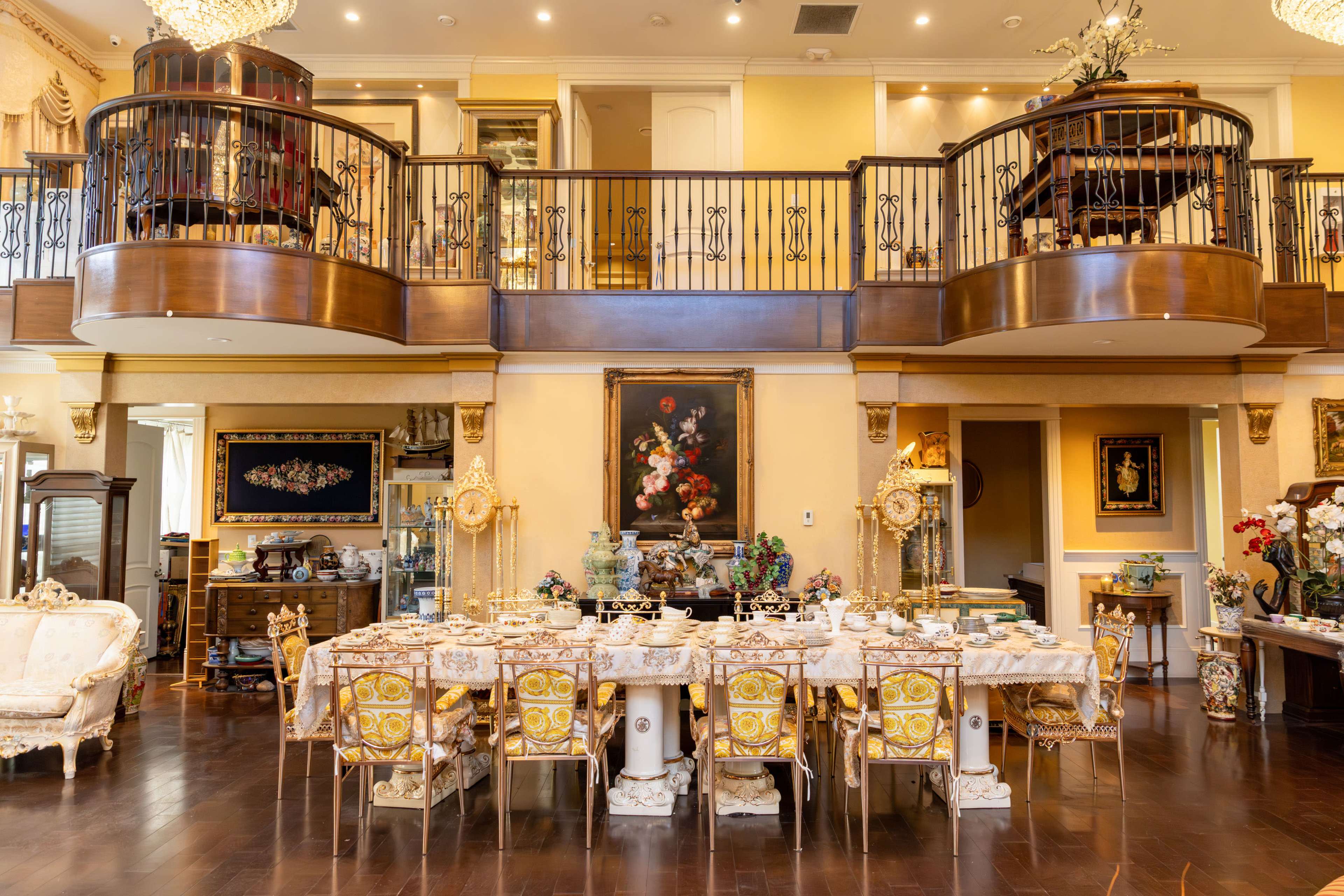 A lavish dining room features an ornate table set for a meal, surrounded by elegant chairs, under a grand chandelier with a decorative balcony above.