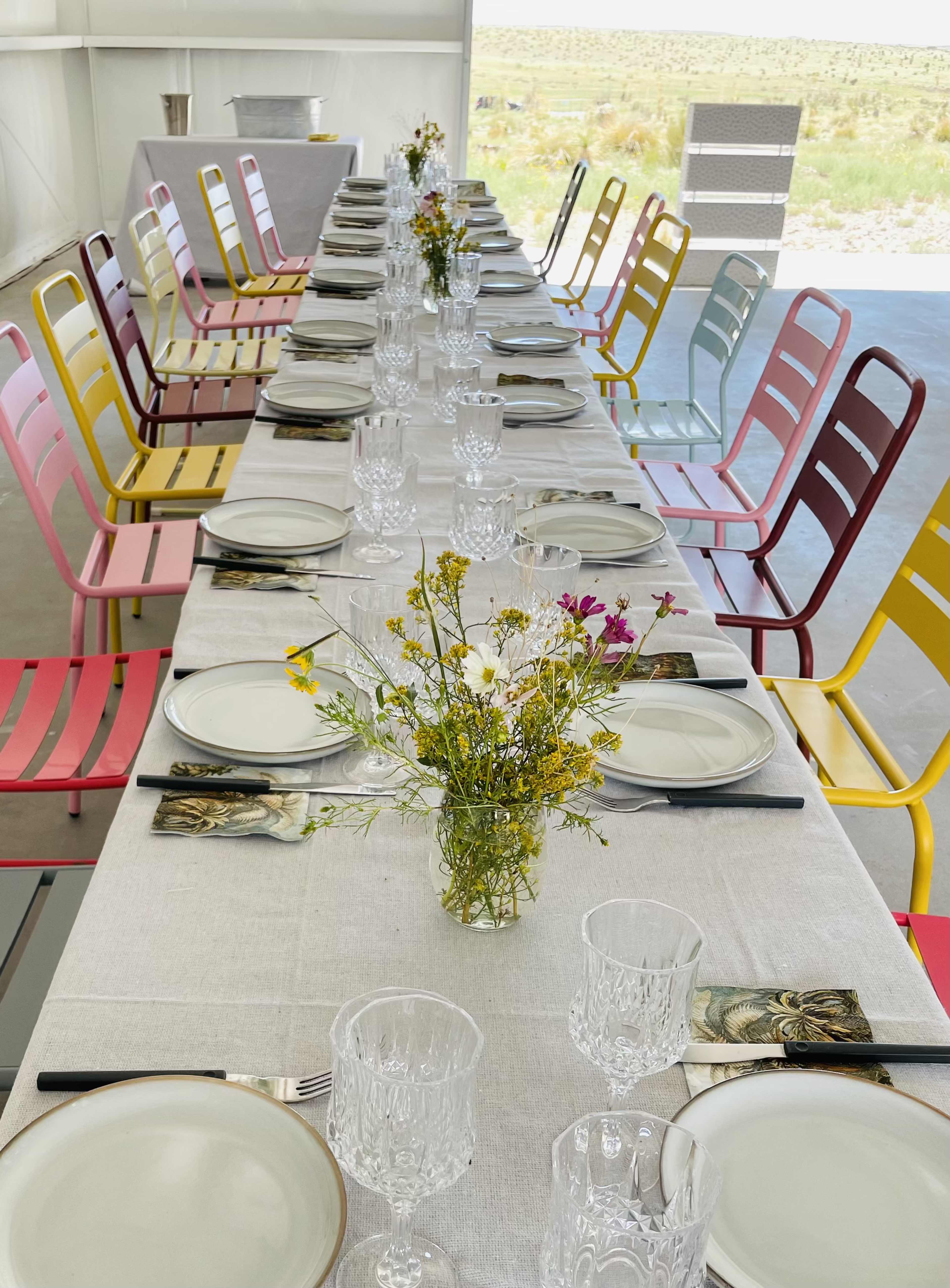 A long dining table is set with plates and glassware, surrounded by colorful chairs and decorated with a small vase of flowers.