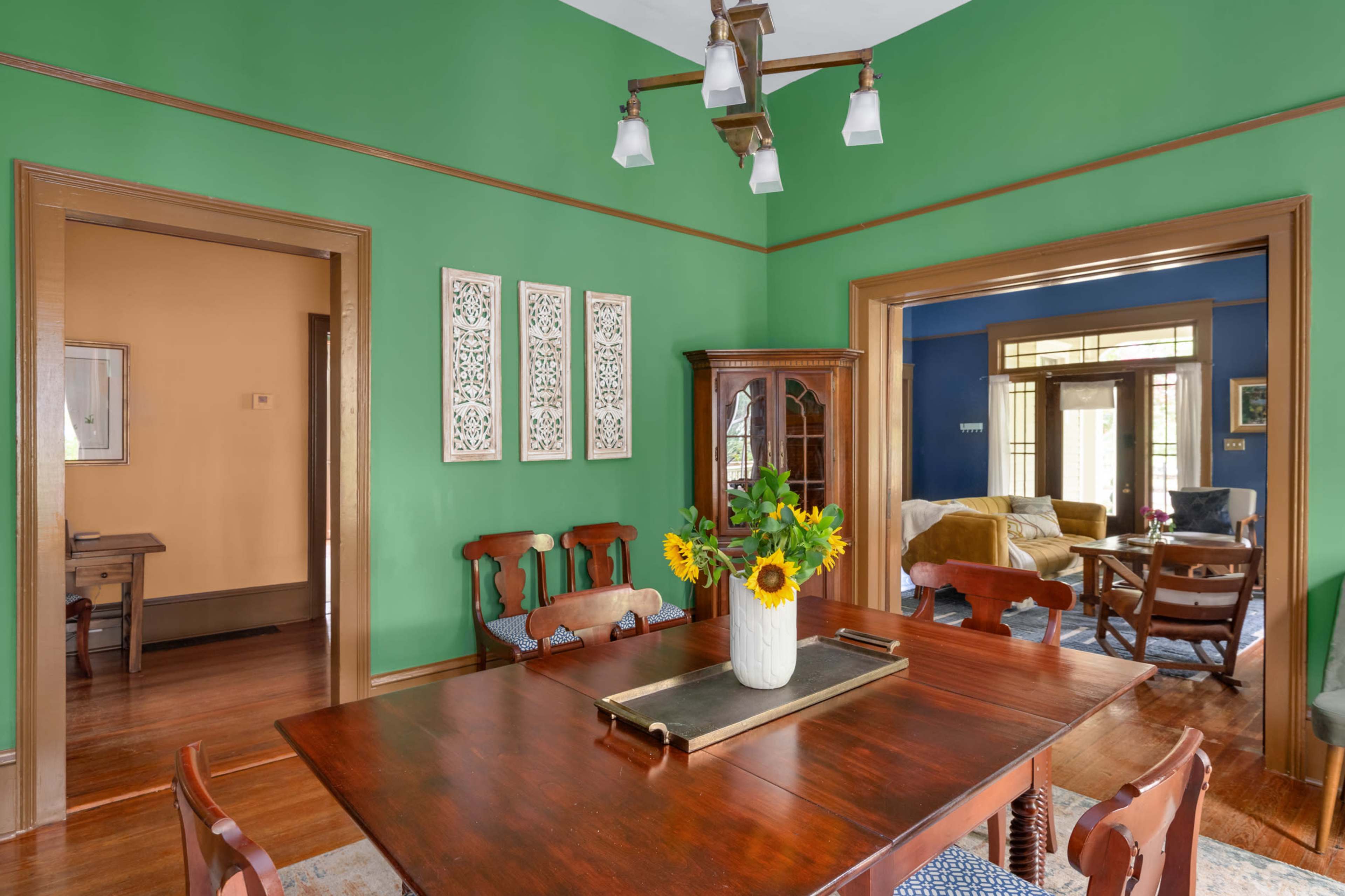 The dining area features a wooden table surrounded by chairs, with green walls and a vase of sunflowers at the center.