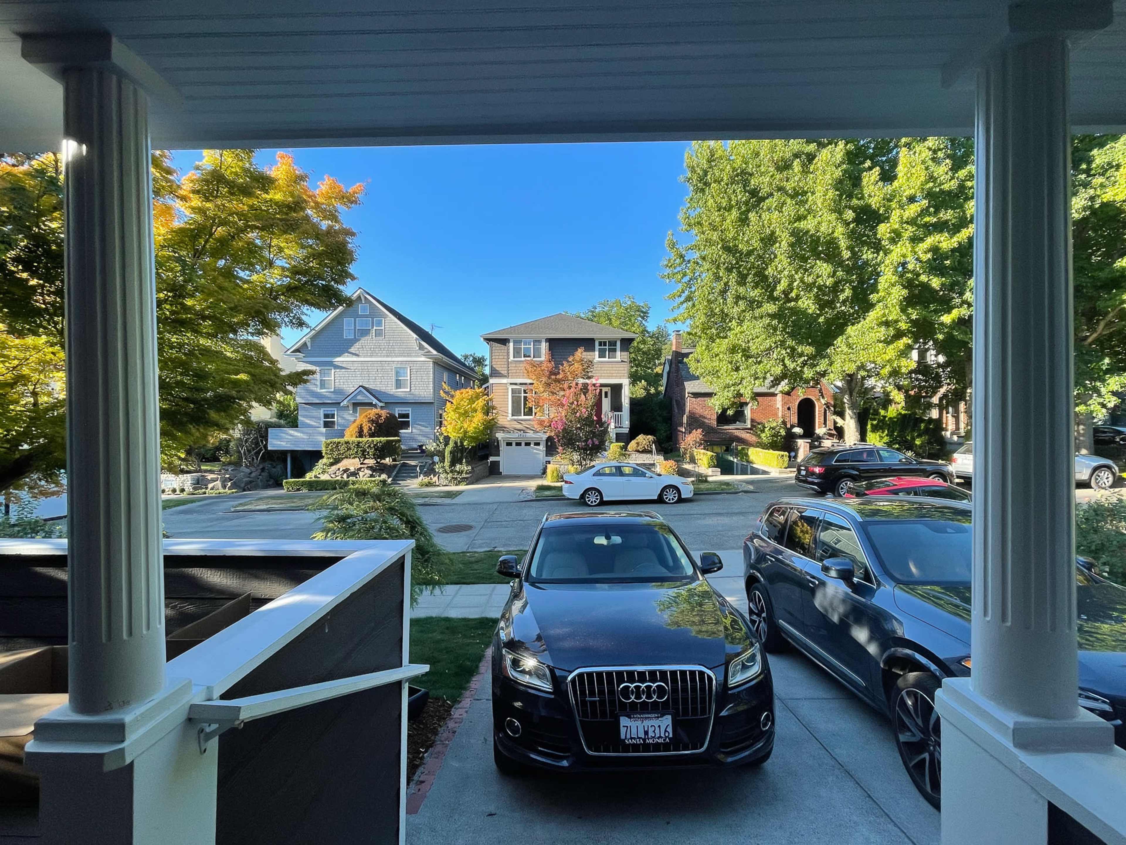A view from a porch overlooking a suburban street with parked cars and houses amidst greenery.