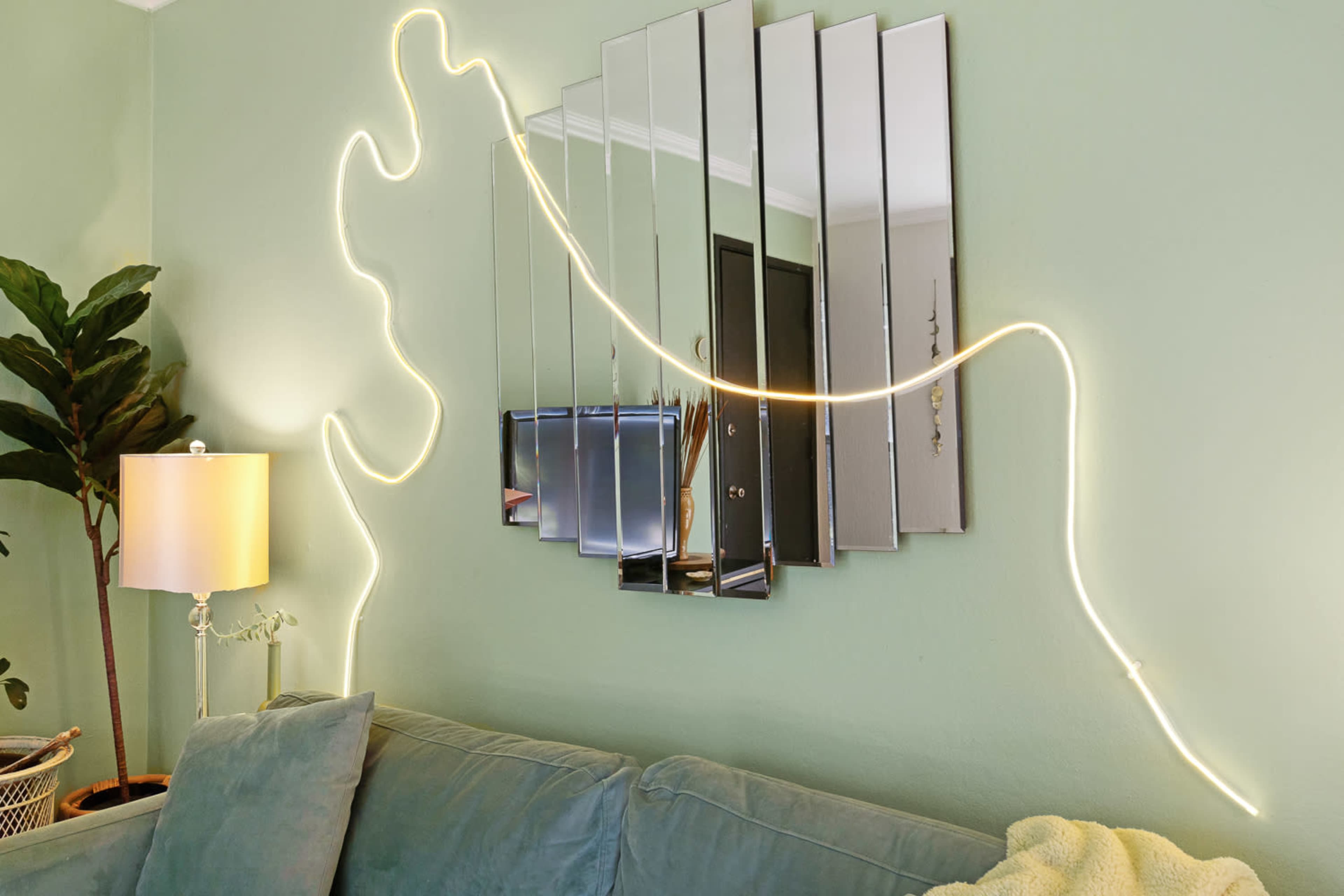 A green living room features a lighted decorative line above a mirrored wall arrangement next to a sofa and a lamp.