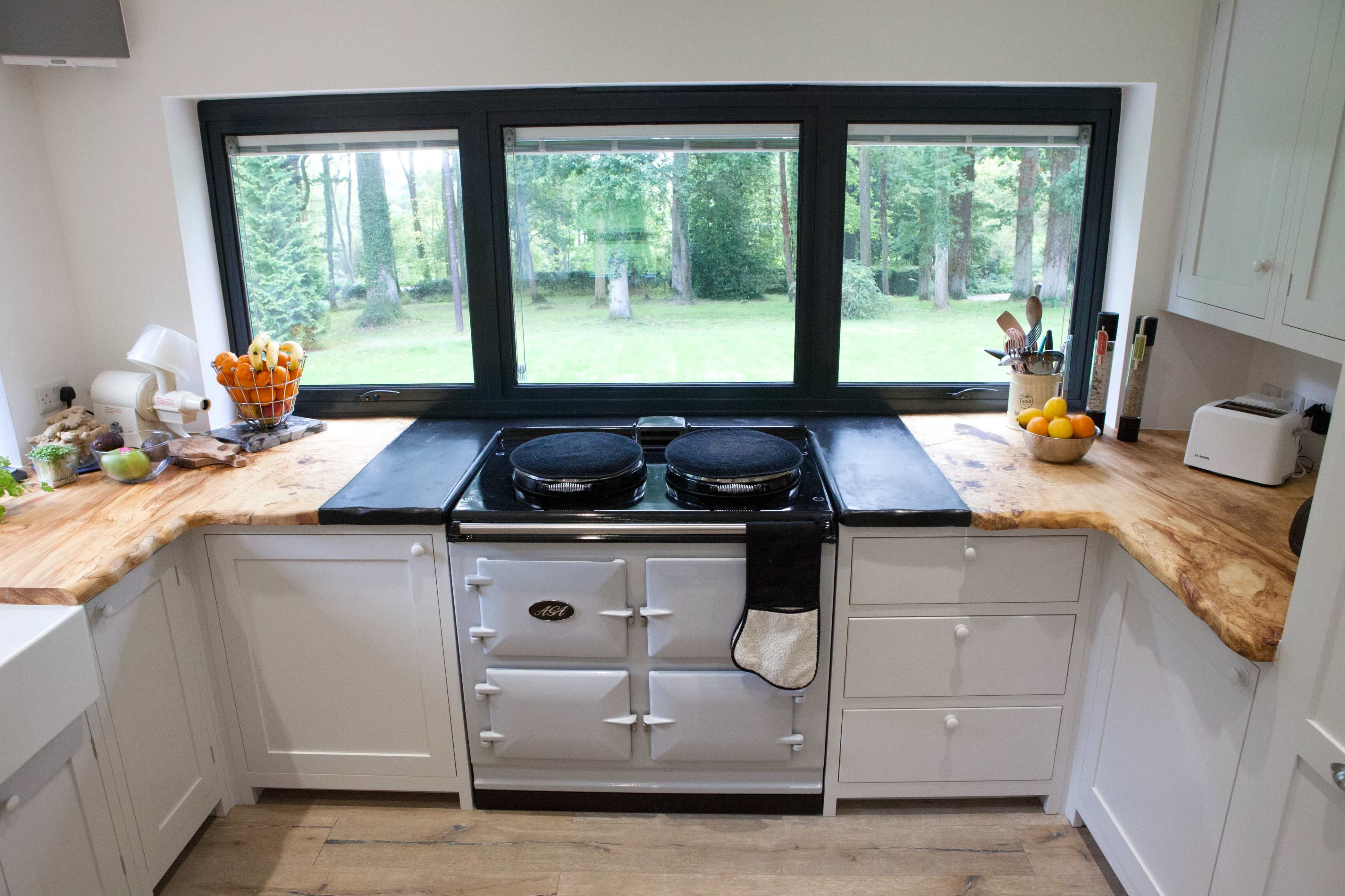 A modern kitchen with a wood countertop and a traditional stove beneath a large window overlooking a green outdoor space.