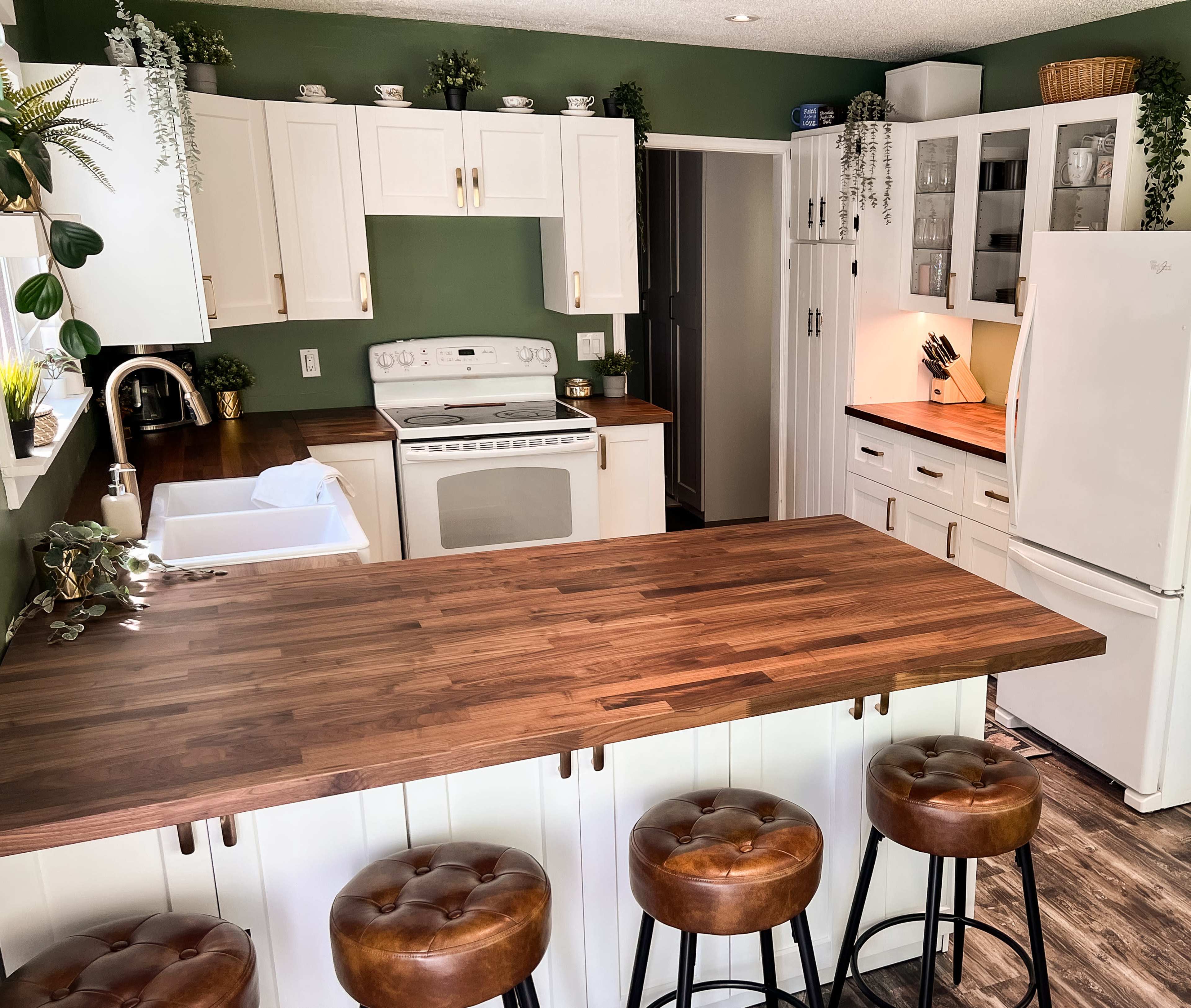 The kitchen features white cabinetry, a wooden countertop island with four barstools, and green walls adorned with plants.