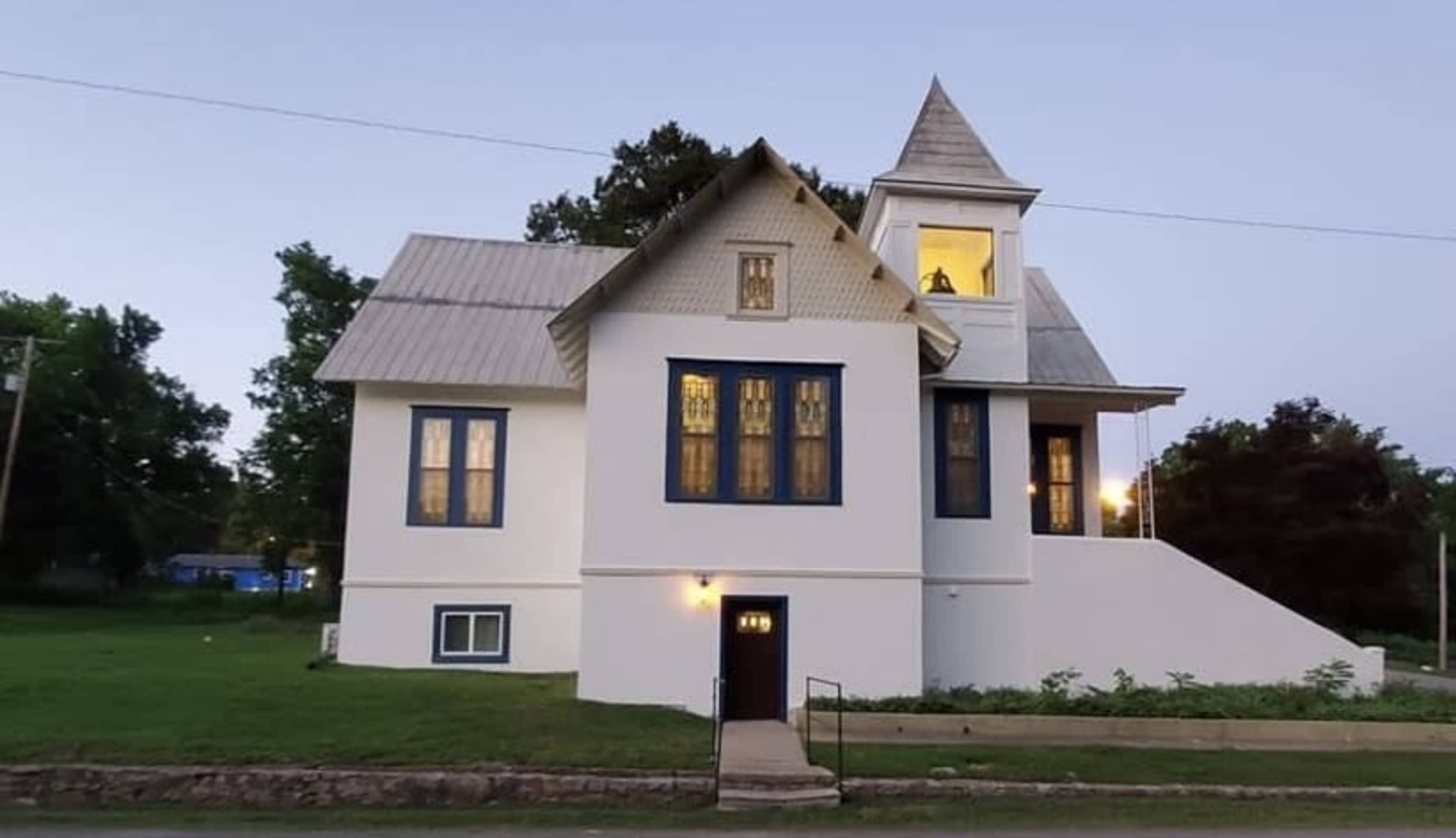 A large, two-story house with a peaked roof and multiple windows stands on a grassy lot, with a well-lit upper room visible at dusk.