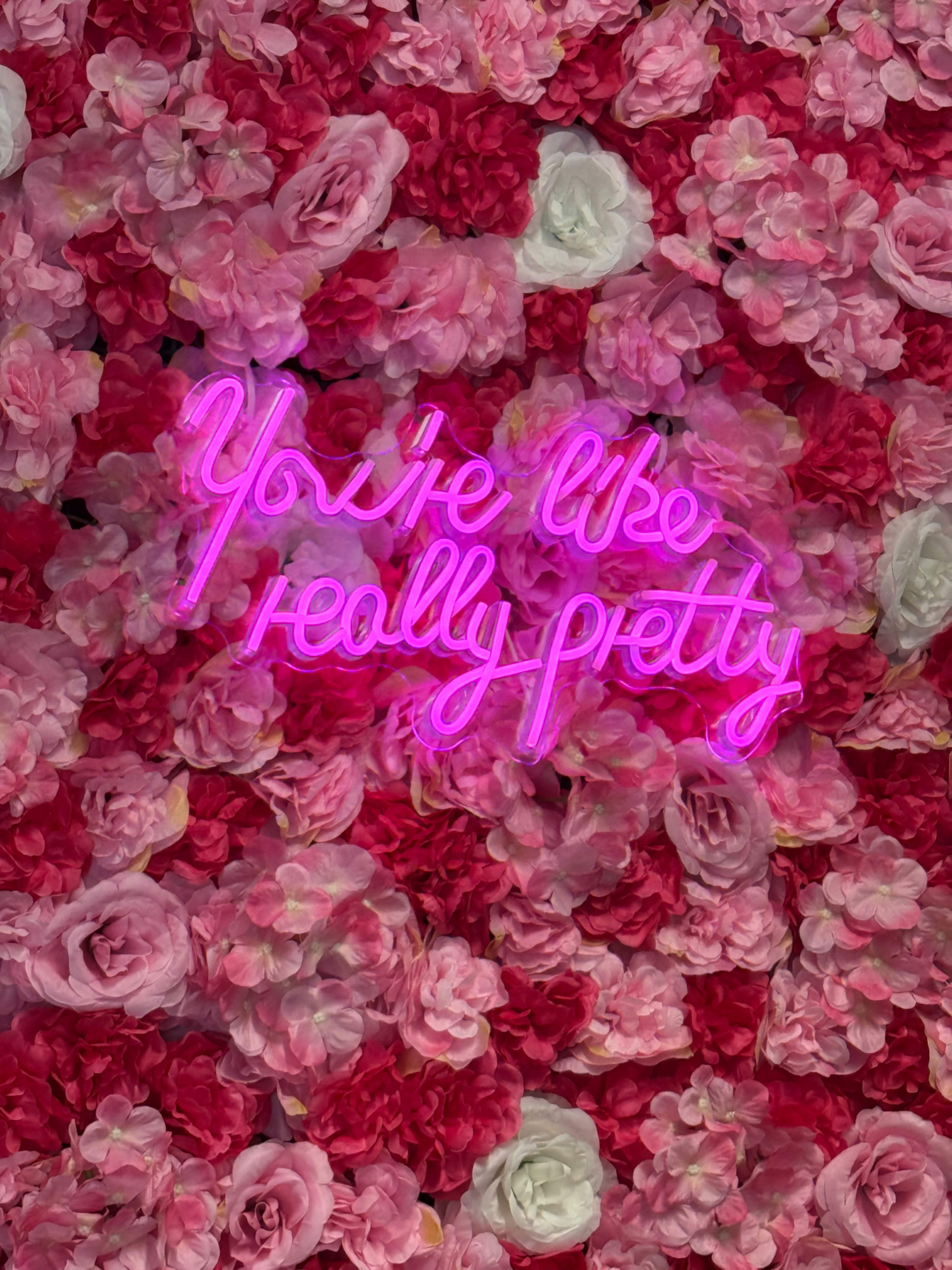 A neon pink sign reading "You're like really pretty" is surrounded by a wall of pink and white artificial flowers.