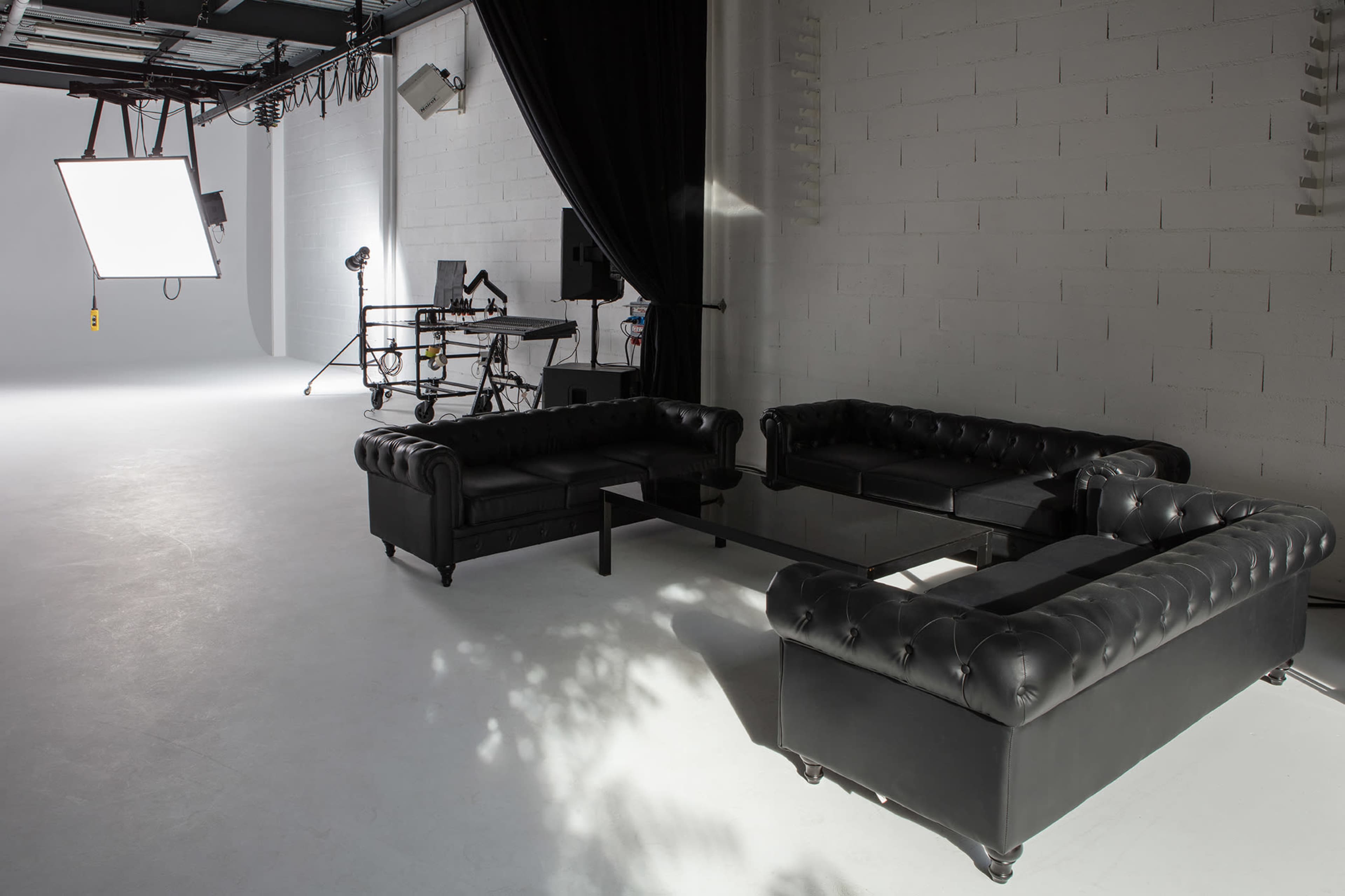 A modern studio setup featuring a black leather sectional couch, a glass coffee table, and various lighting equipment in a minimalist space.