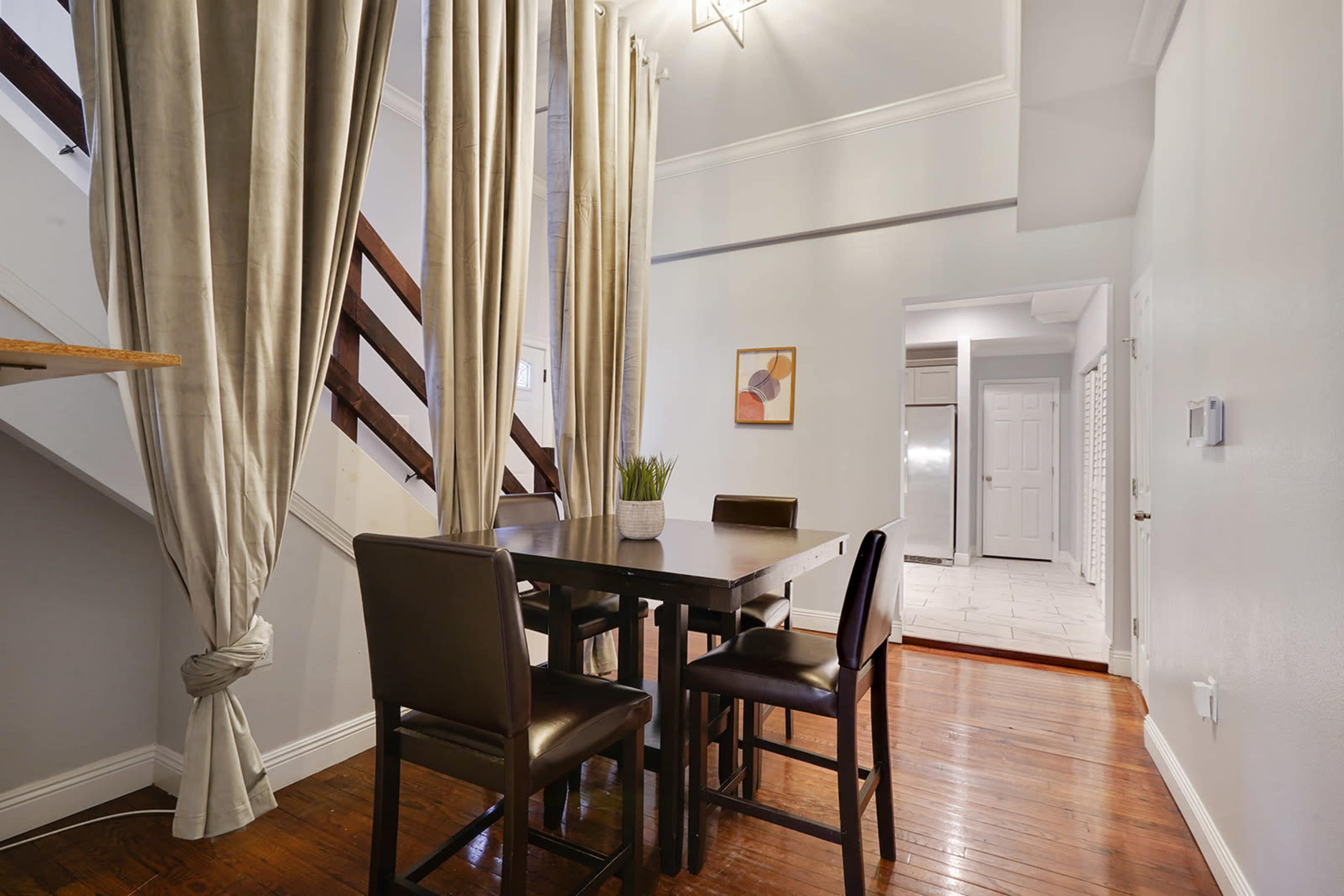 A dining area features a wooden table with four chairs, flanked by curtains and surrounded by a staircase and an open doorway leading to a kitchen.