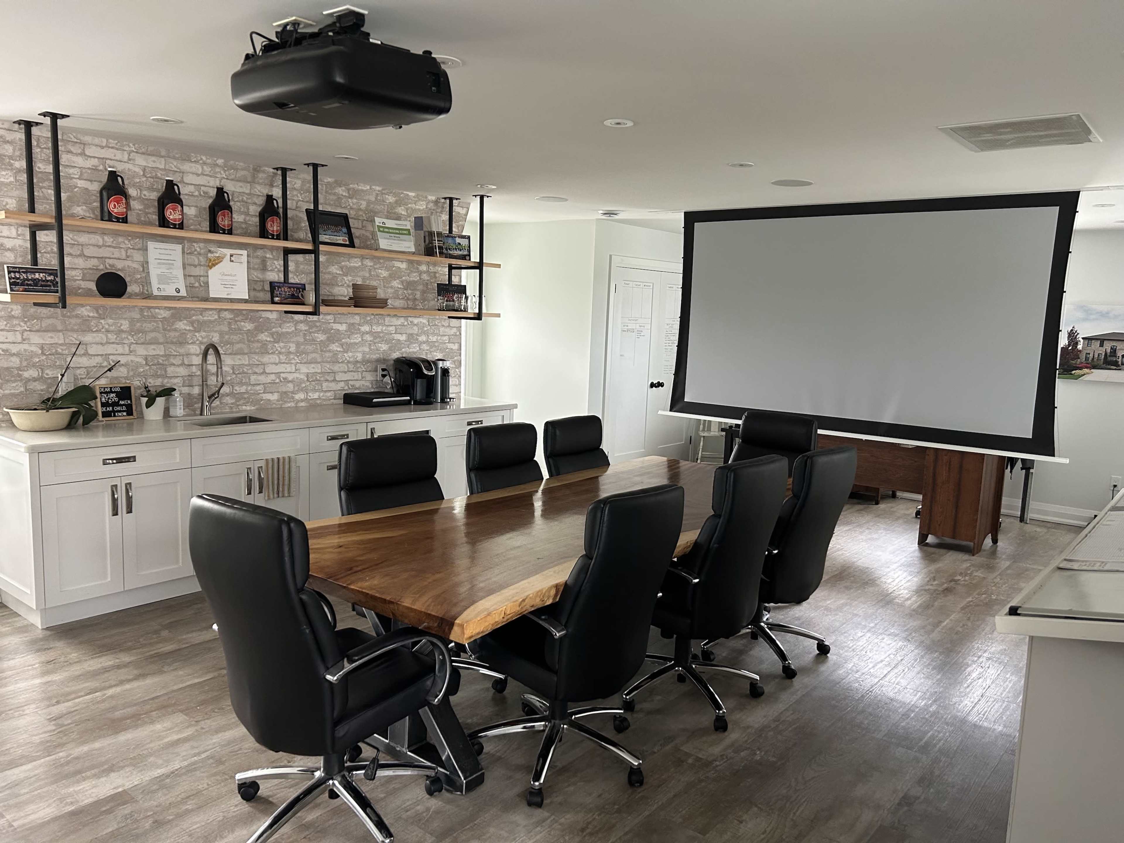 A modern conference room featuring a large wooden table surrounded by black office chairs, a projection screen, and shelves displaying various items.
