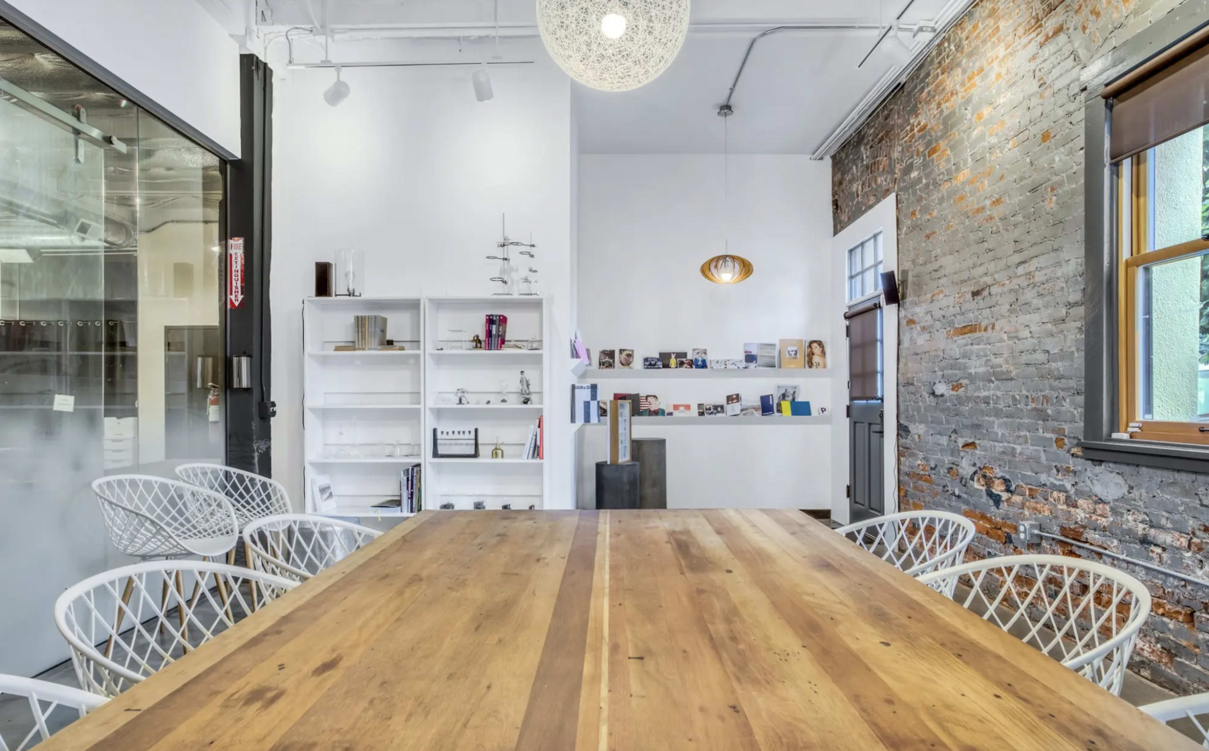 The image shows a modern meeting room featuring a large wooden table, white chairs, exposed brick walls, and a display shelf against a white wall.