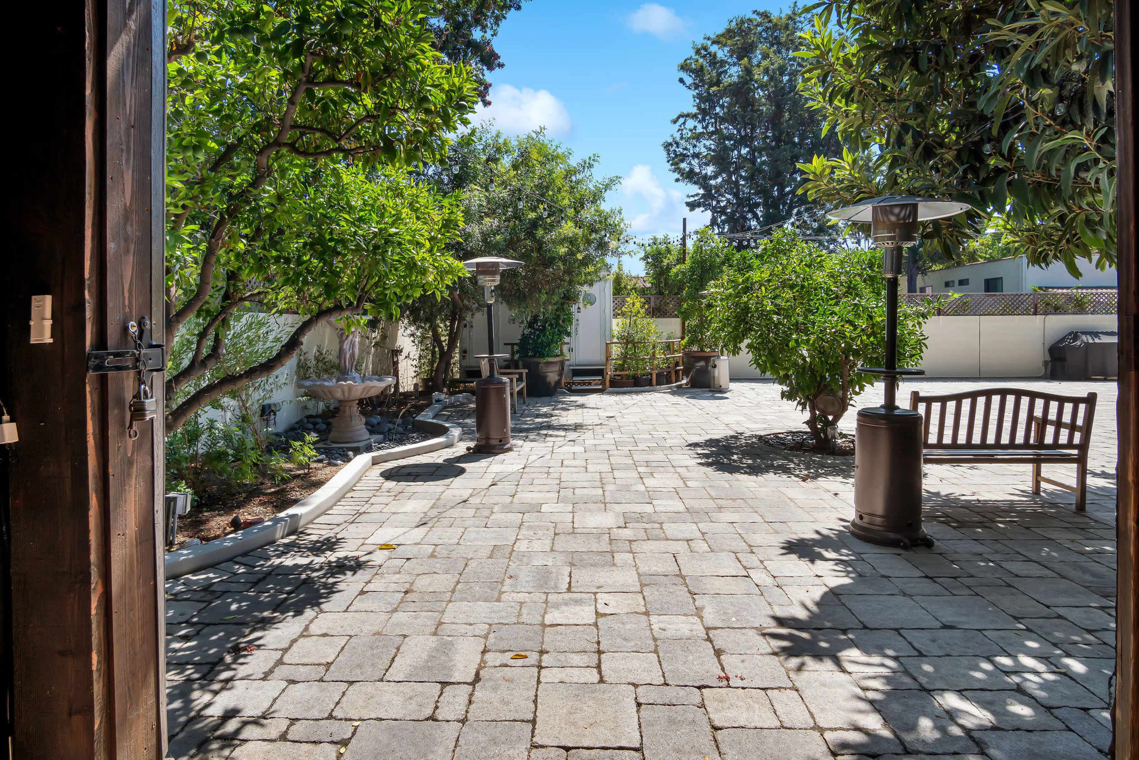 The image shows a paved outdoor courtyard with trees, a bench, and patio heaters, set against a clear blue sky.