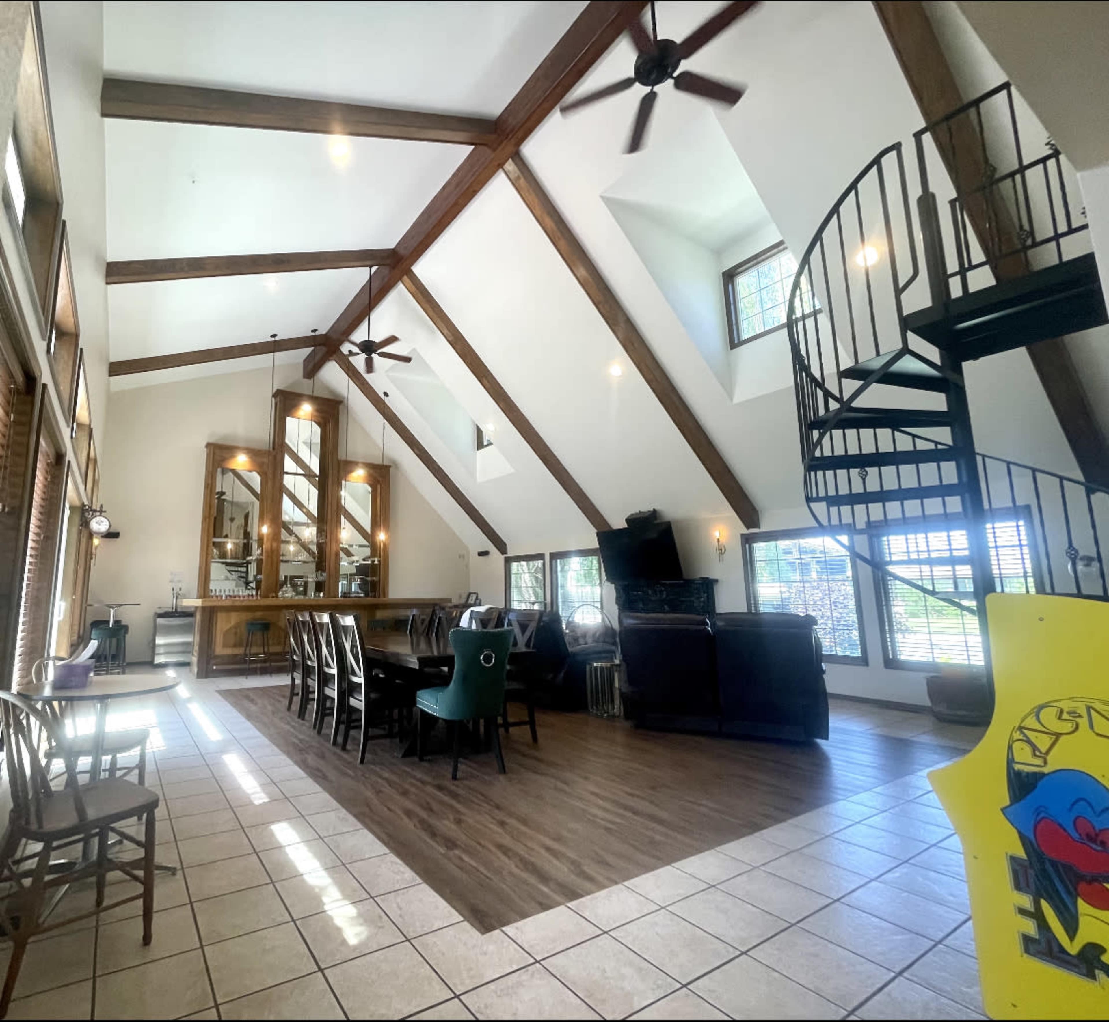 A spacious living area with high ceilings, exposed wooden beams, a dining table, a staircase, and large windows allowing natural light.