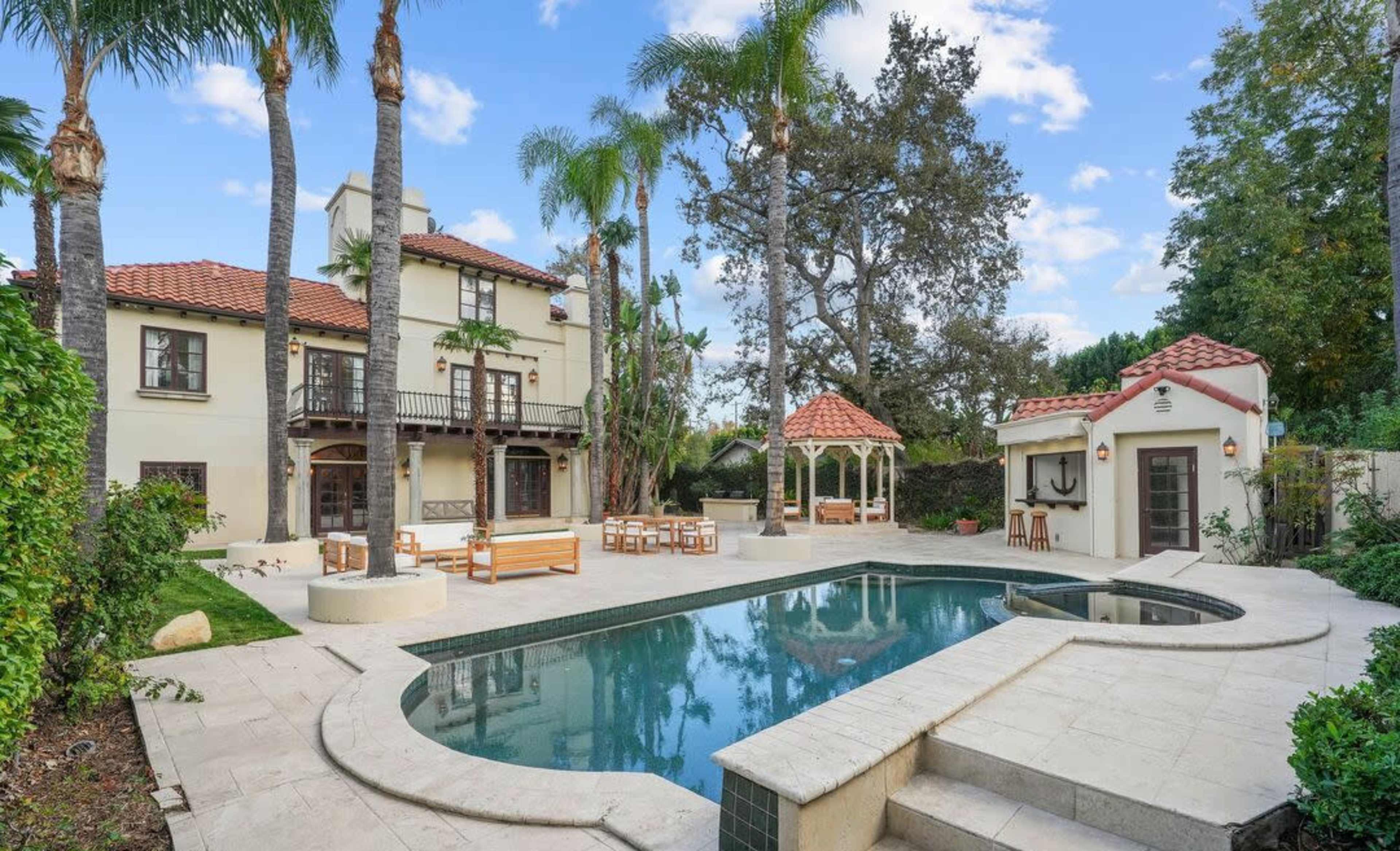 The image shows a spacious outdoor area featuring a rectangular swimming pool surrounded by palm trees, a gazebo, and a large multi-story house with a red-tiled roof.