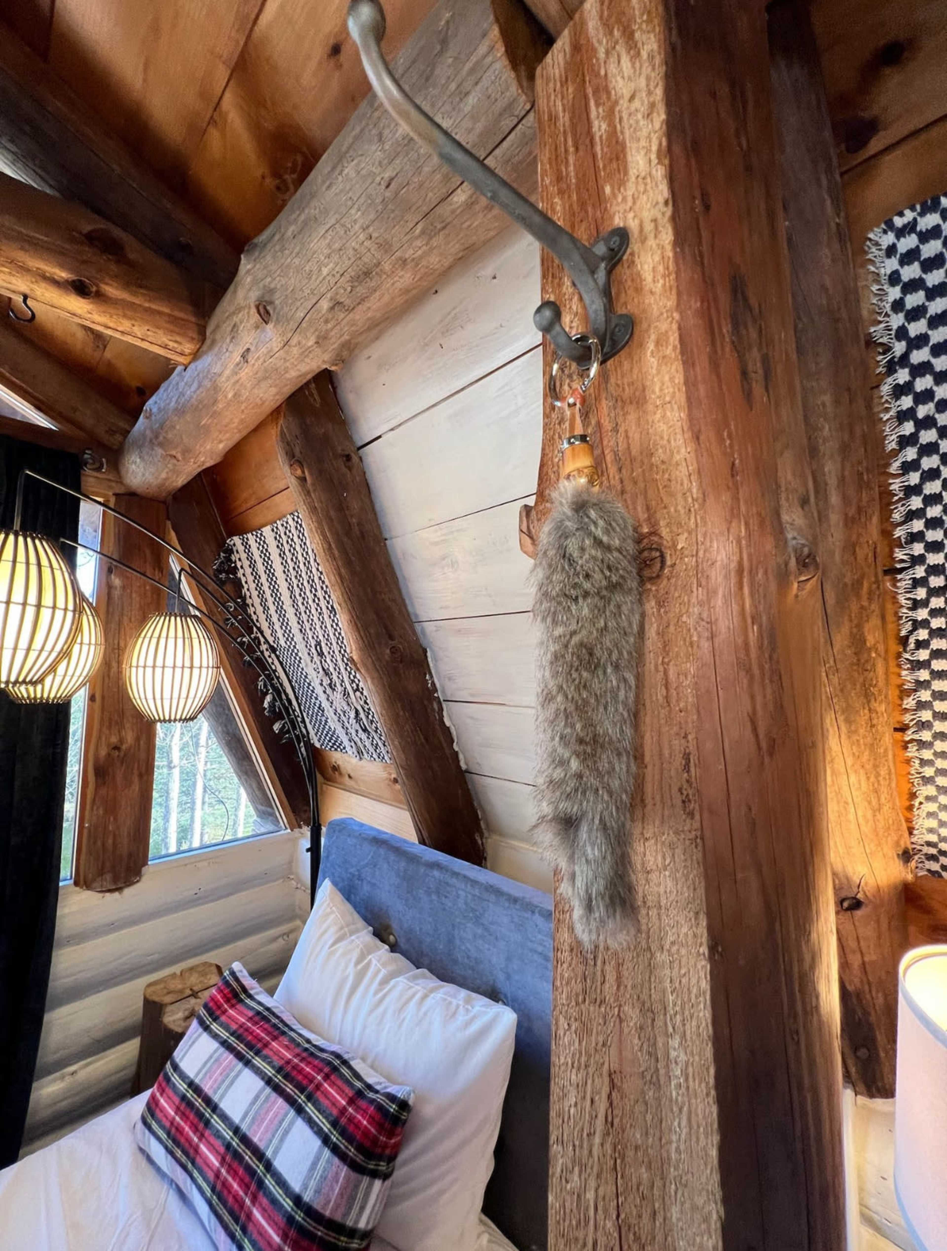 The image shows a rustic bedroom with wooden beams, a bed with plaid bedding, and a fur accessory hanging from a metal hook on a wooden post.