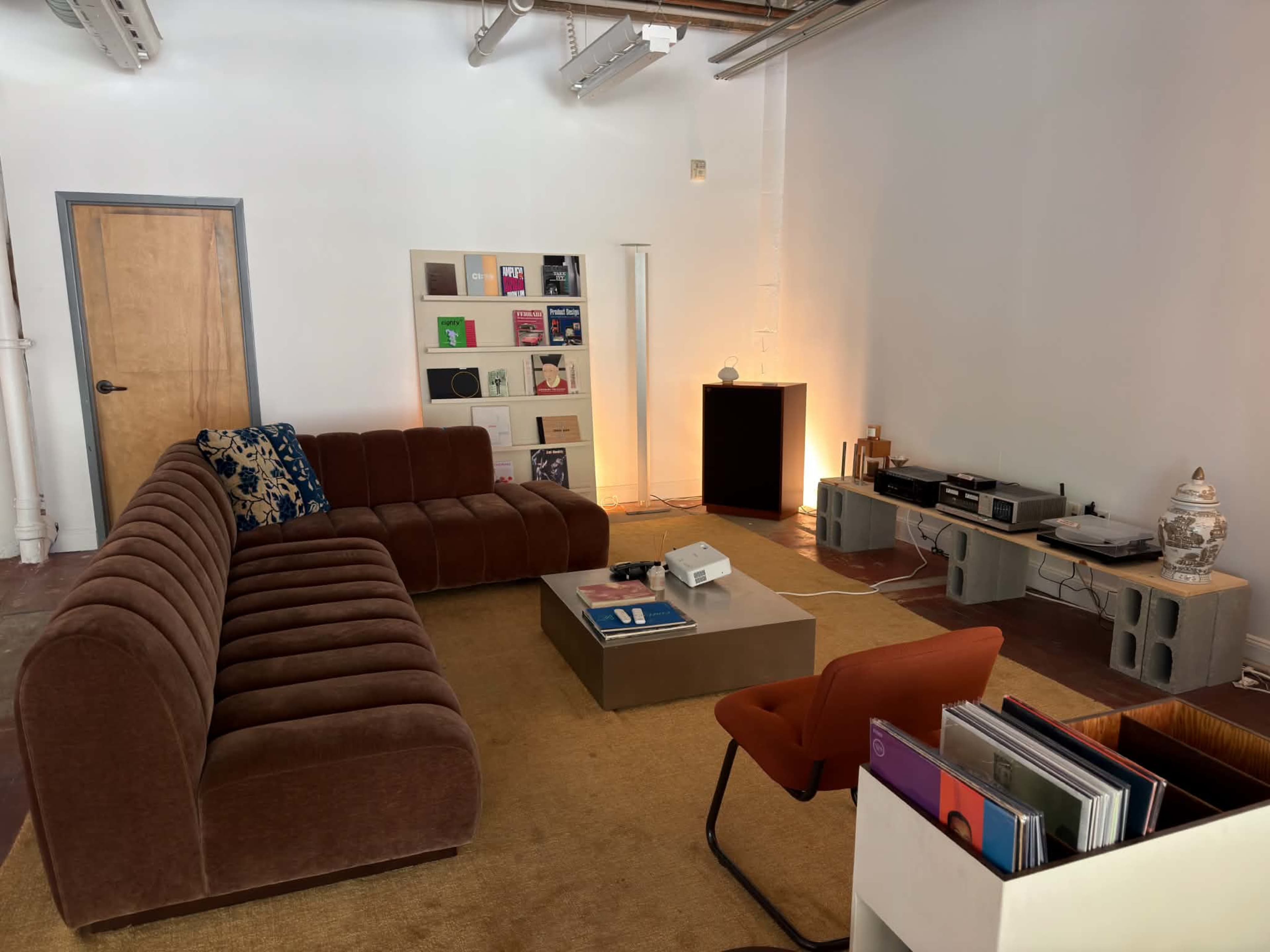 The image shows a modern living room with a brown sectional sofa, a coffee table, a record player setup, and shelves displaying vinyl records.