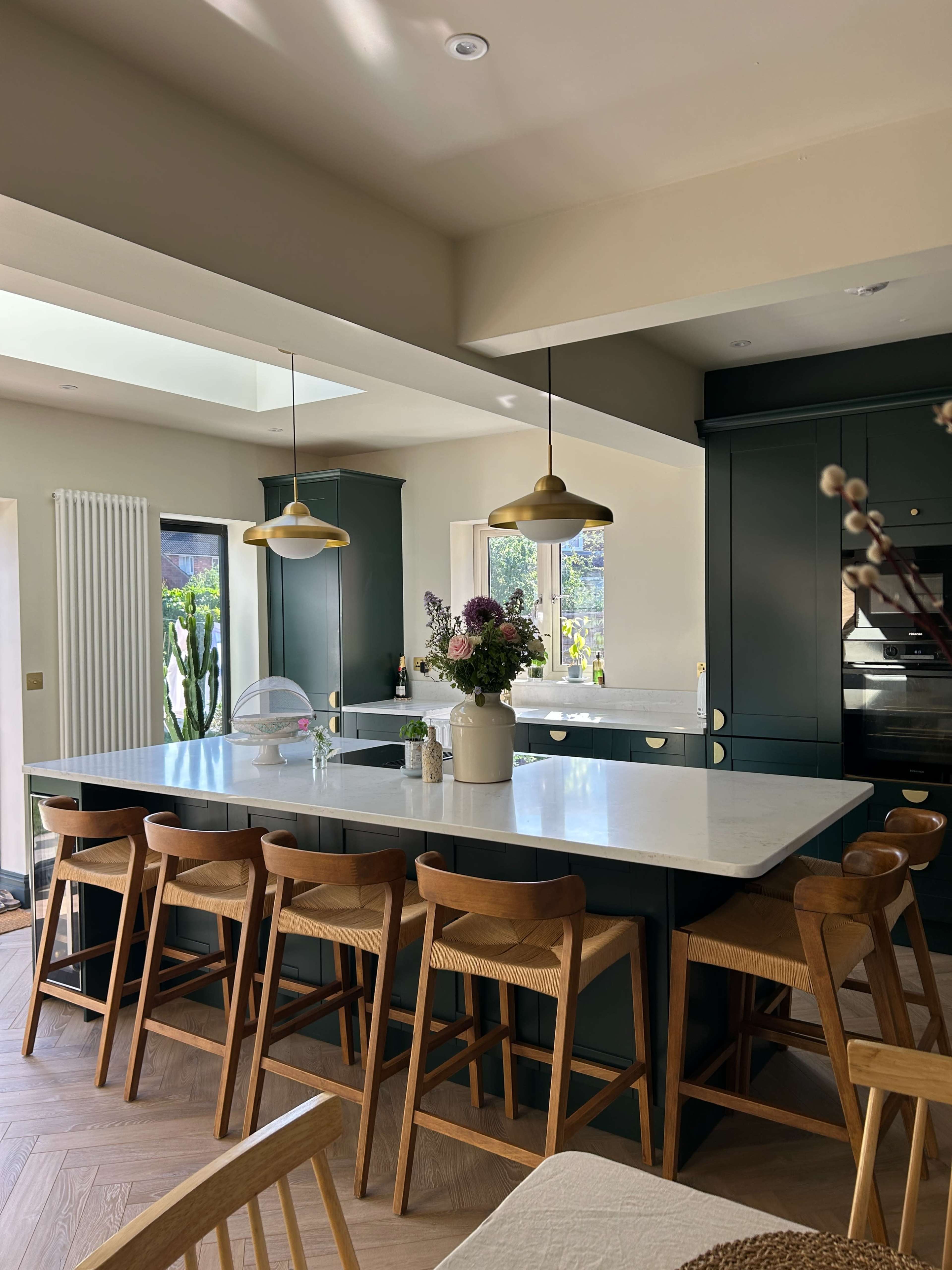 The image shows a modern kitchen with a large island, wooden bar stools, and green cabinetry, complemented by natural light streaming through large windows.