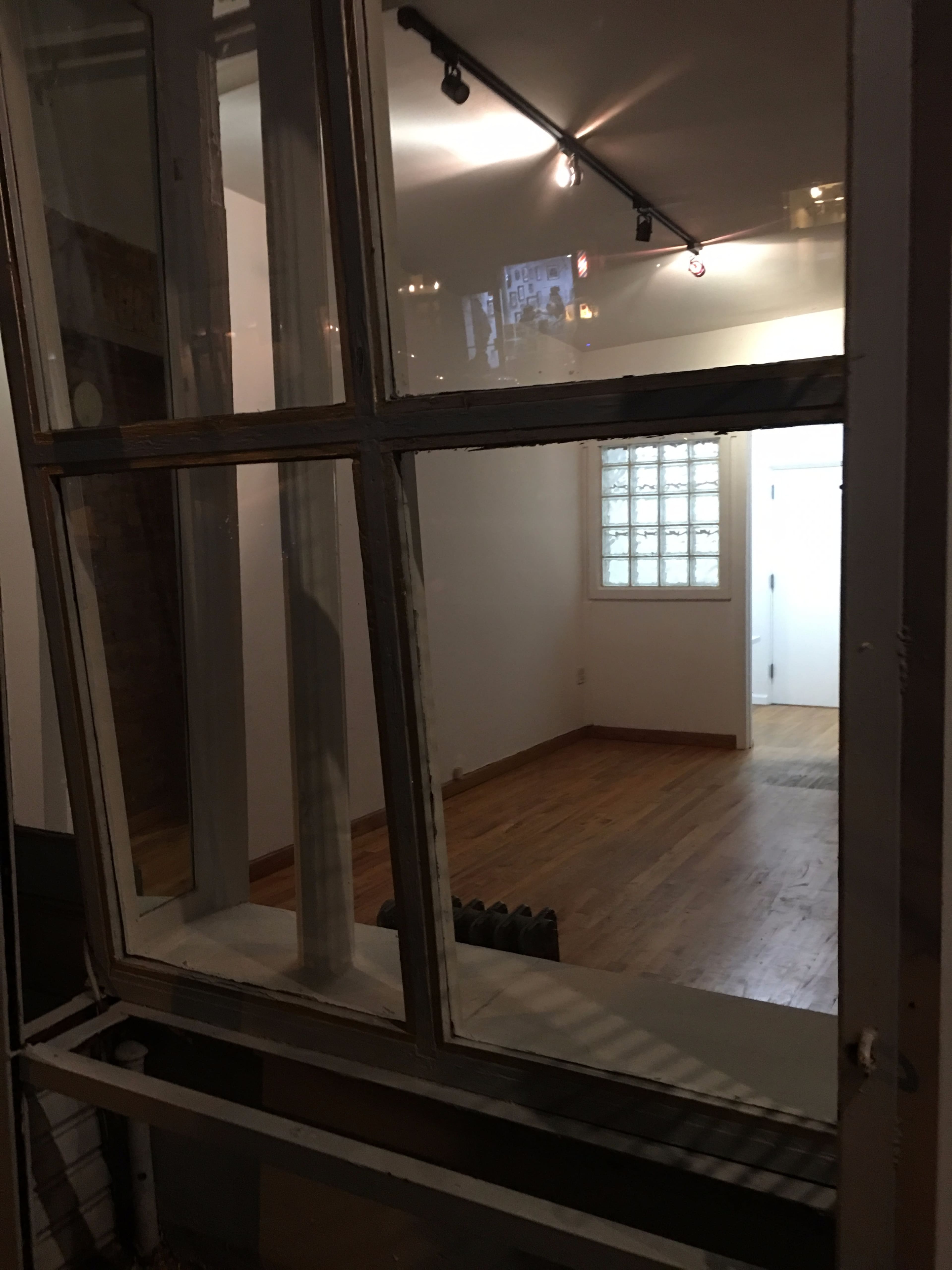 The image shows a partially open window framing a view into an empty room with wooden flooring and glass block windows.