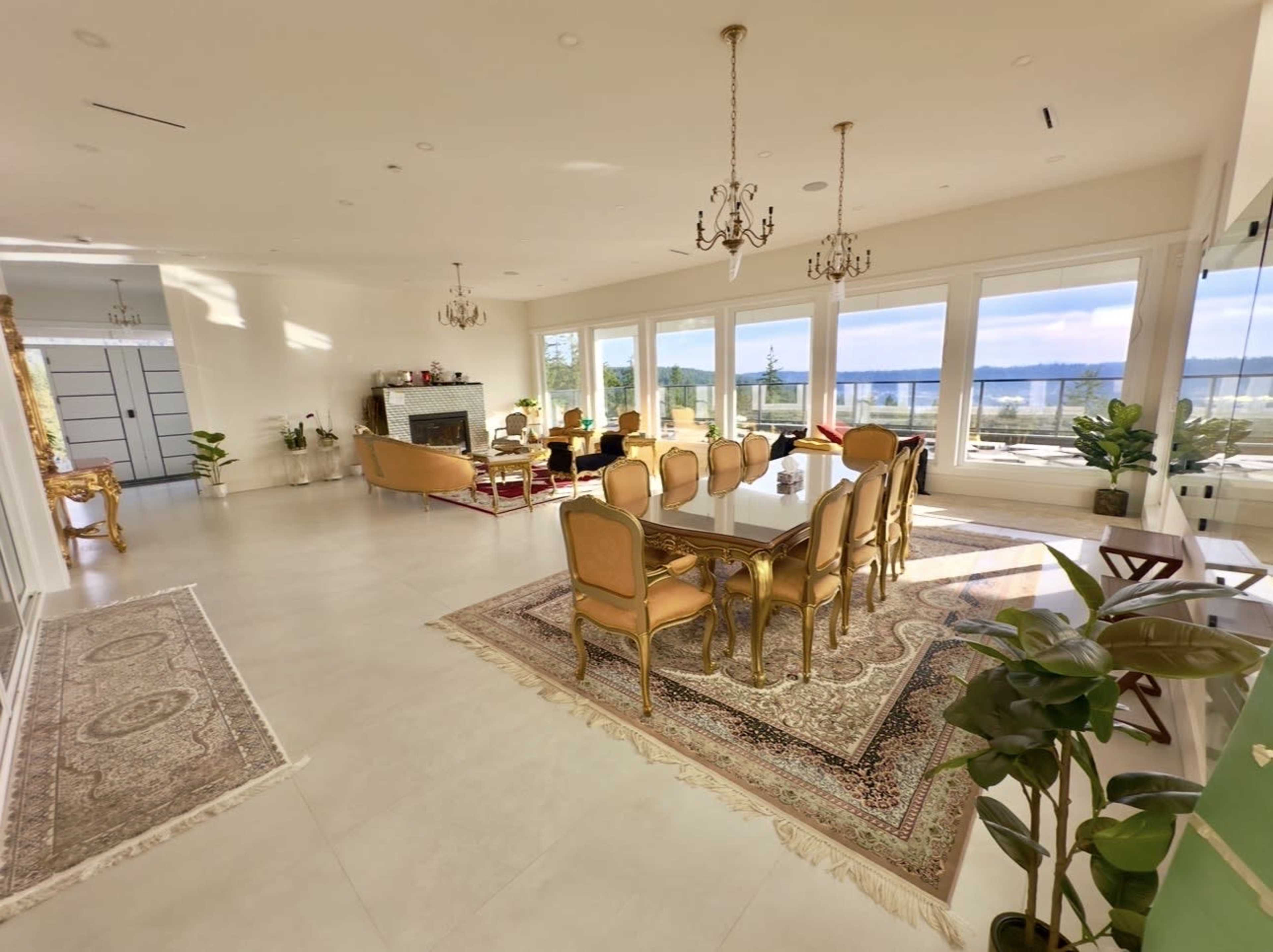 The image depicts a spacious and brightly lit living area with a dining table and ornate chairs, large windows showing a view of the outdoors, and decorative plants.