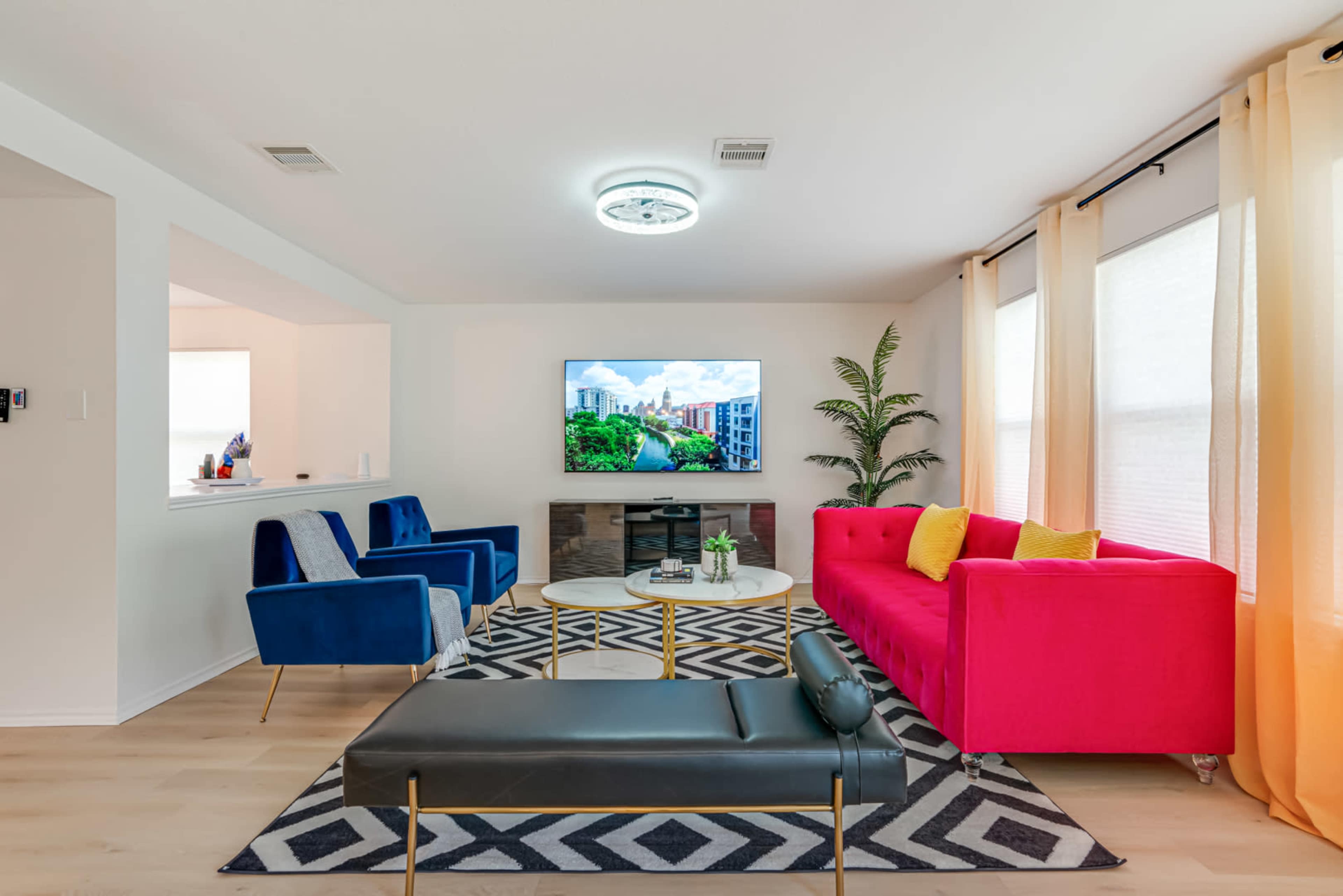 A modern living room featuring two blue chairs, a red sofa, a black ottoman, a round coffee table, and a television mounted on the wall.