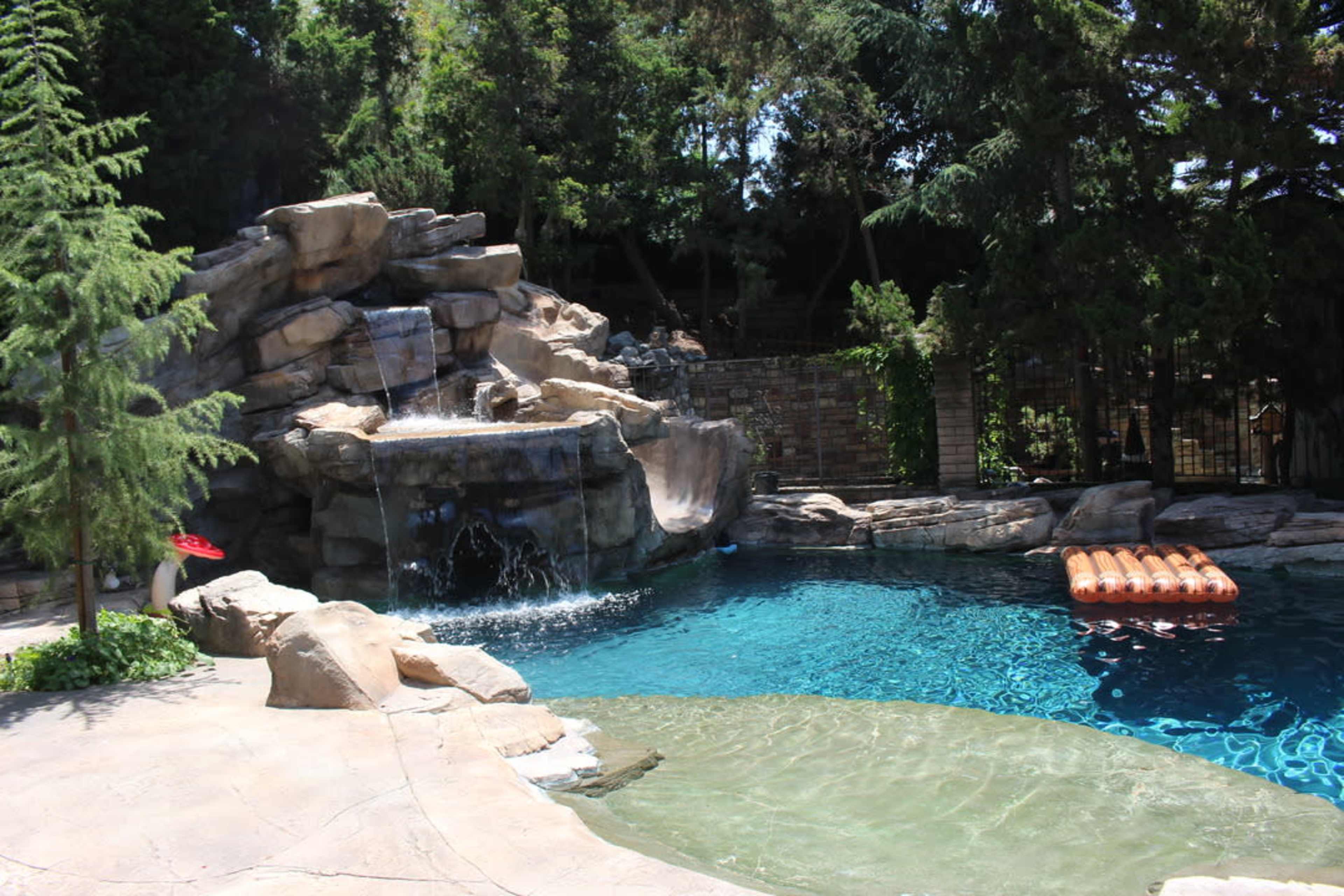 The image shows a swimming pool with a rock formation and waterfall on one side, surrounded by greenery and outdoor features.