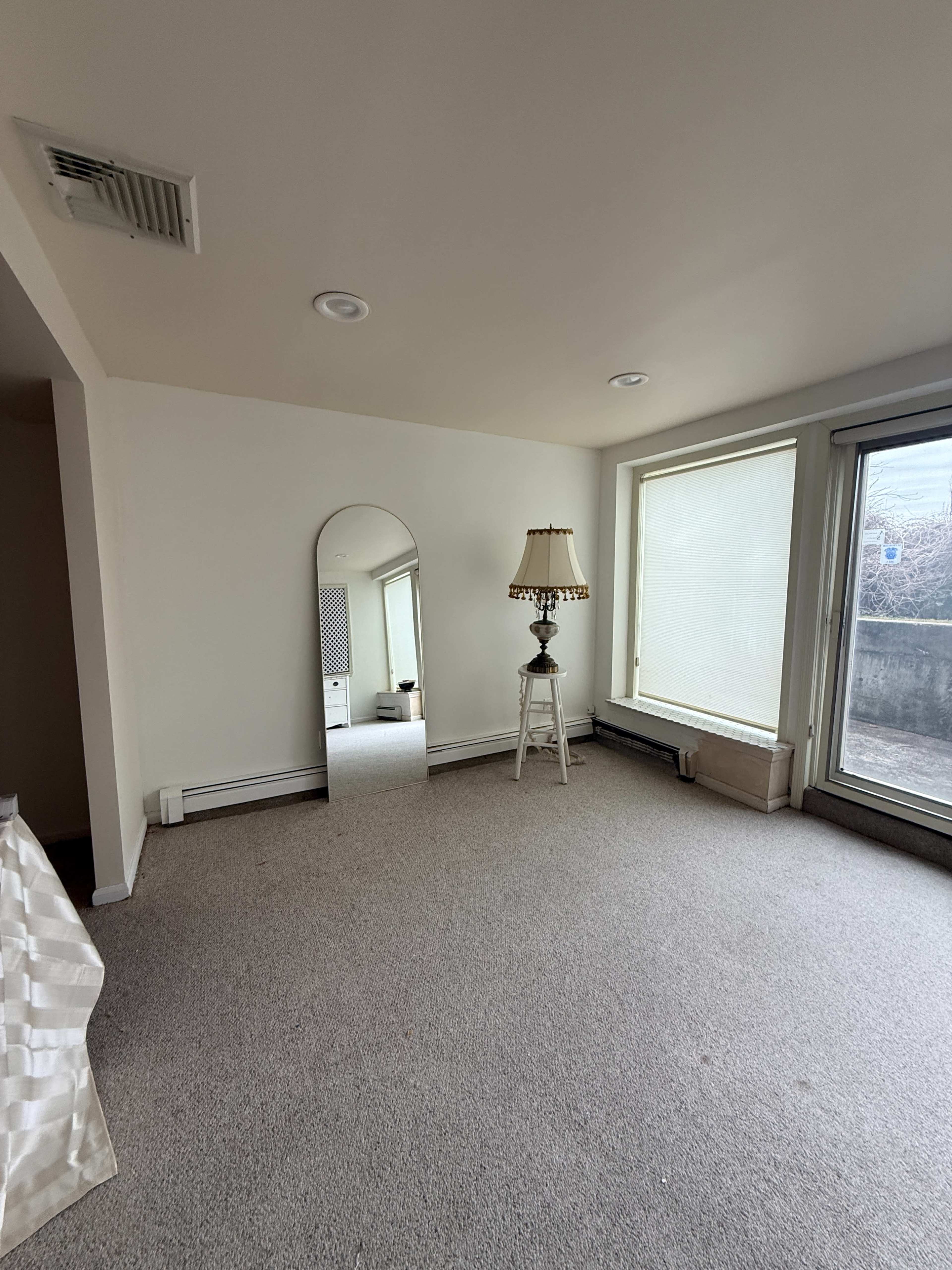 A spacious, empty room features a large mirror, a lamp on a small table, and two windows allowing natural light.