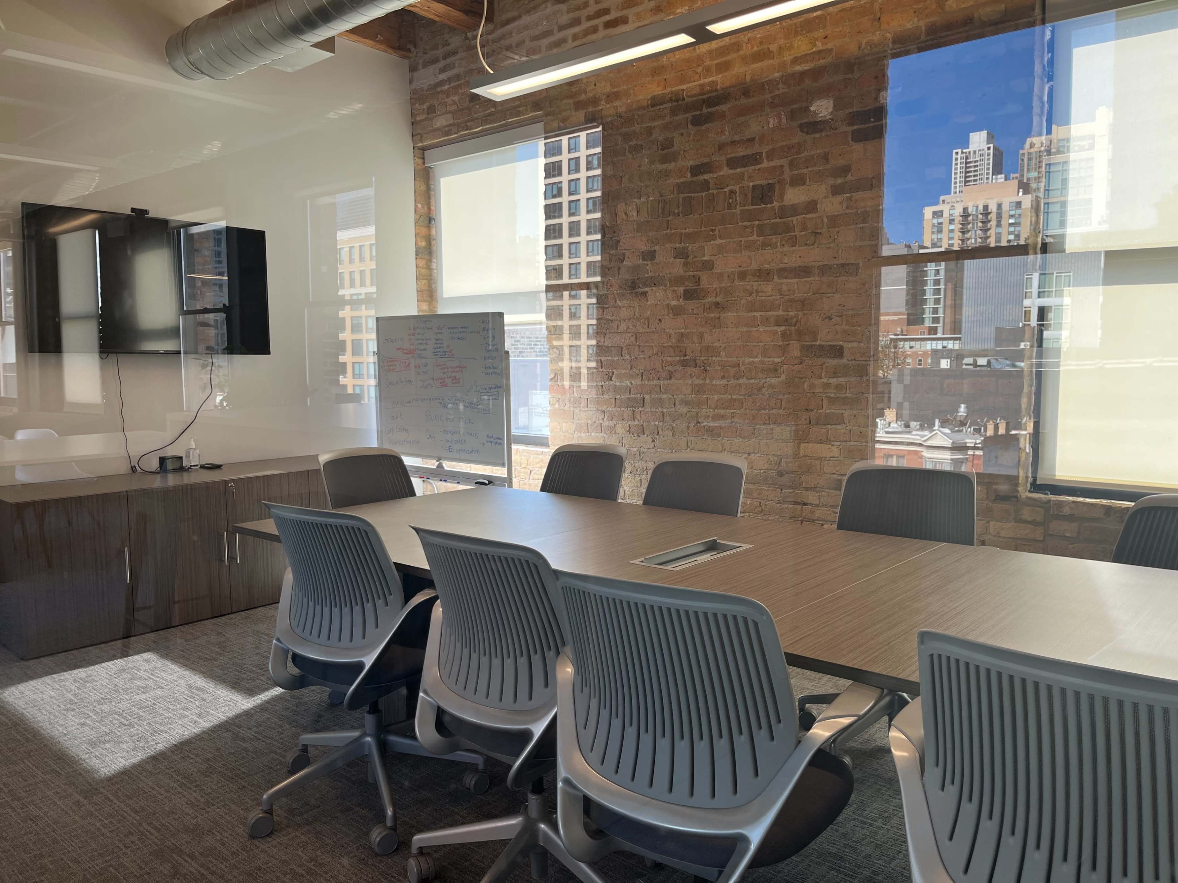 A modern conference room features a large rectangular table surrounded by gray chairs, with a whiteboard and a wall-mounted screen visible in a well-lit space with large windows.