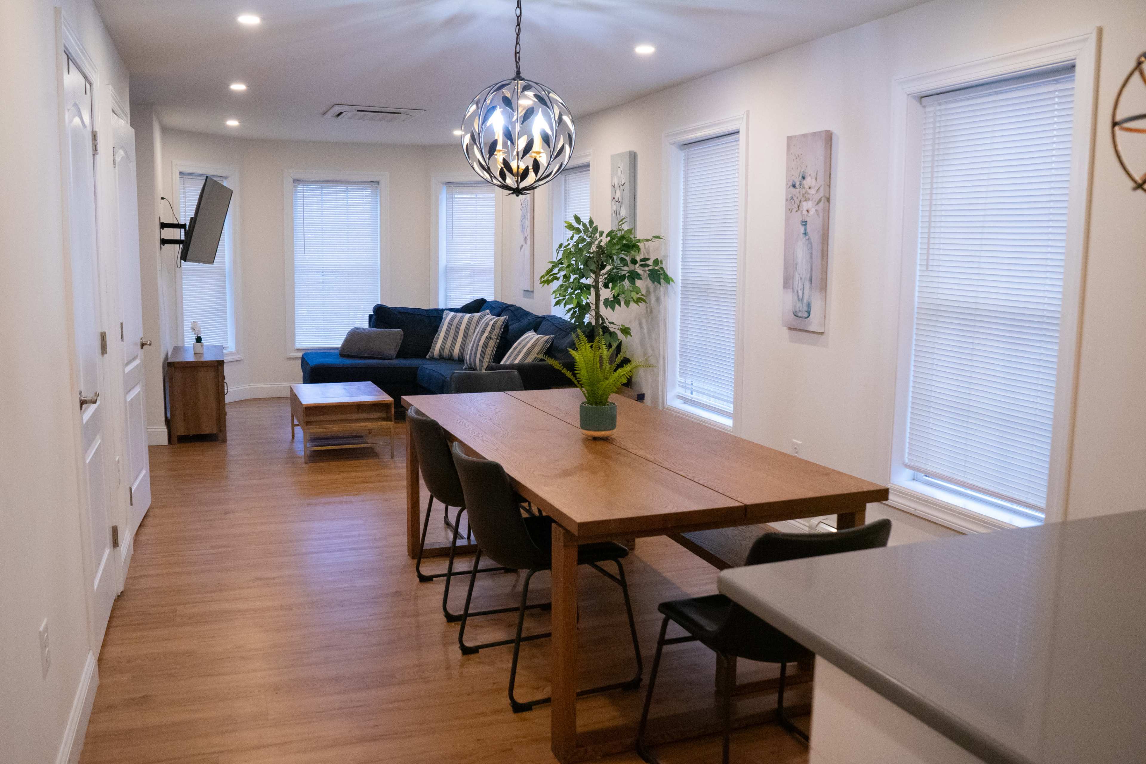 A modern living space features a dining table, chairs, a sofa, and large windows with natural light.