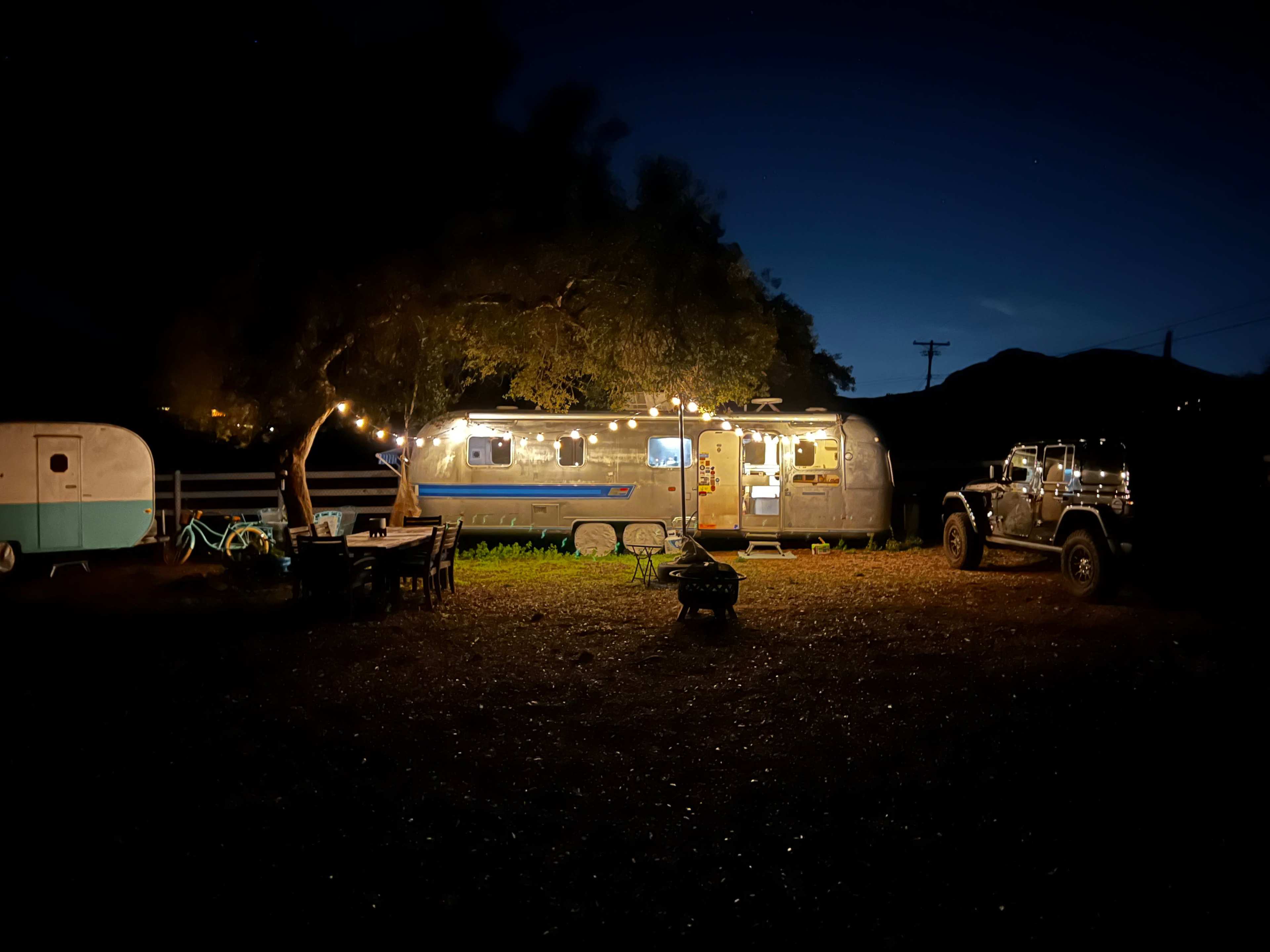 An Airstream trailer is illuminated at night with string lights, surrounded by outdoor seating and a Jeep nearby.