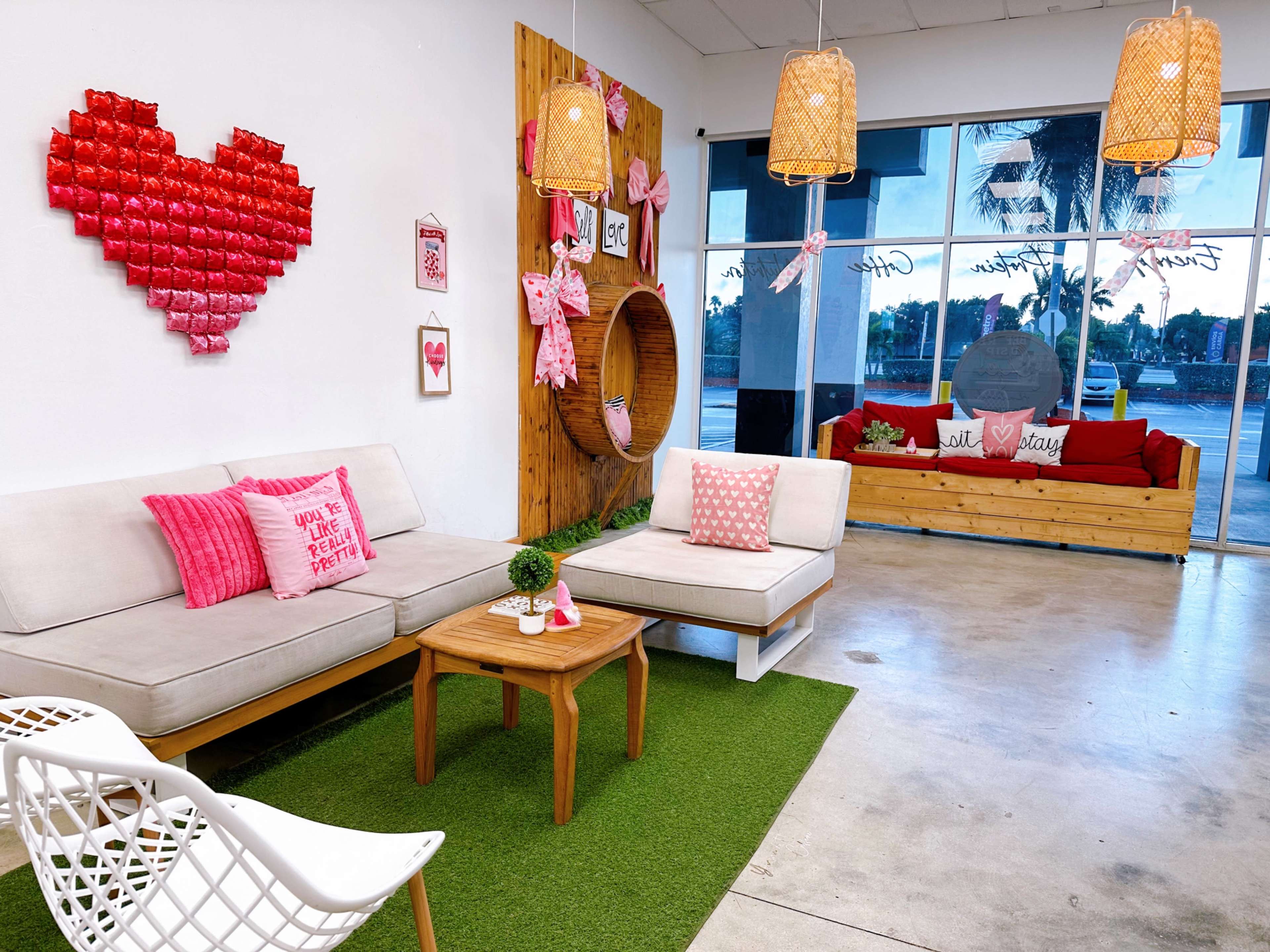 The image shows a cozy seating area decorated with heart-themed decor, including a wall with red heart-shaped items and various cushions, along with wooden furniture and green grass-like flooring.