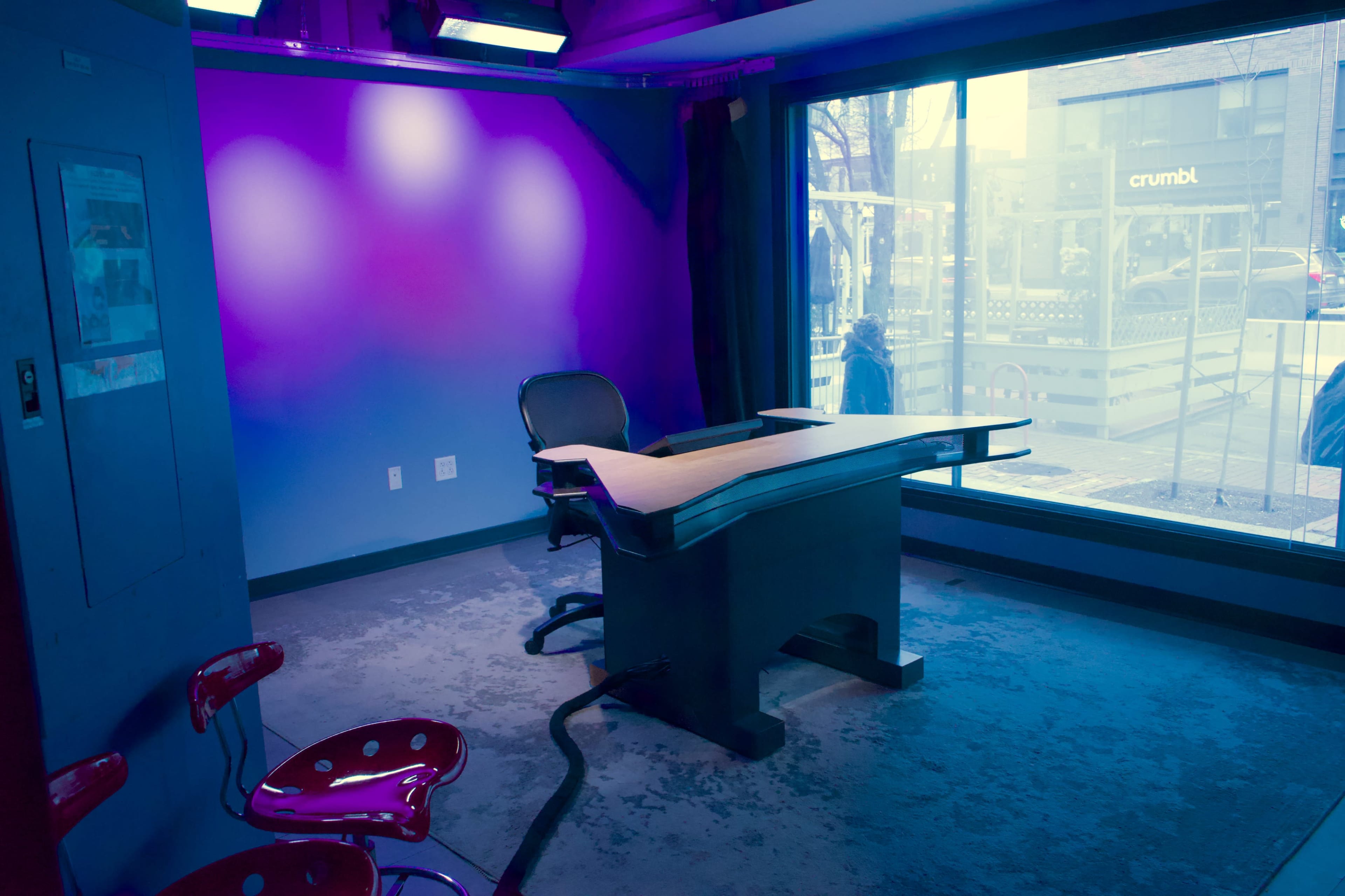 A modern studio space features a desk and chairs, with large windows displaying a city street outside.