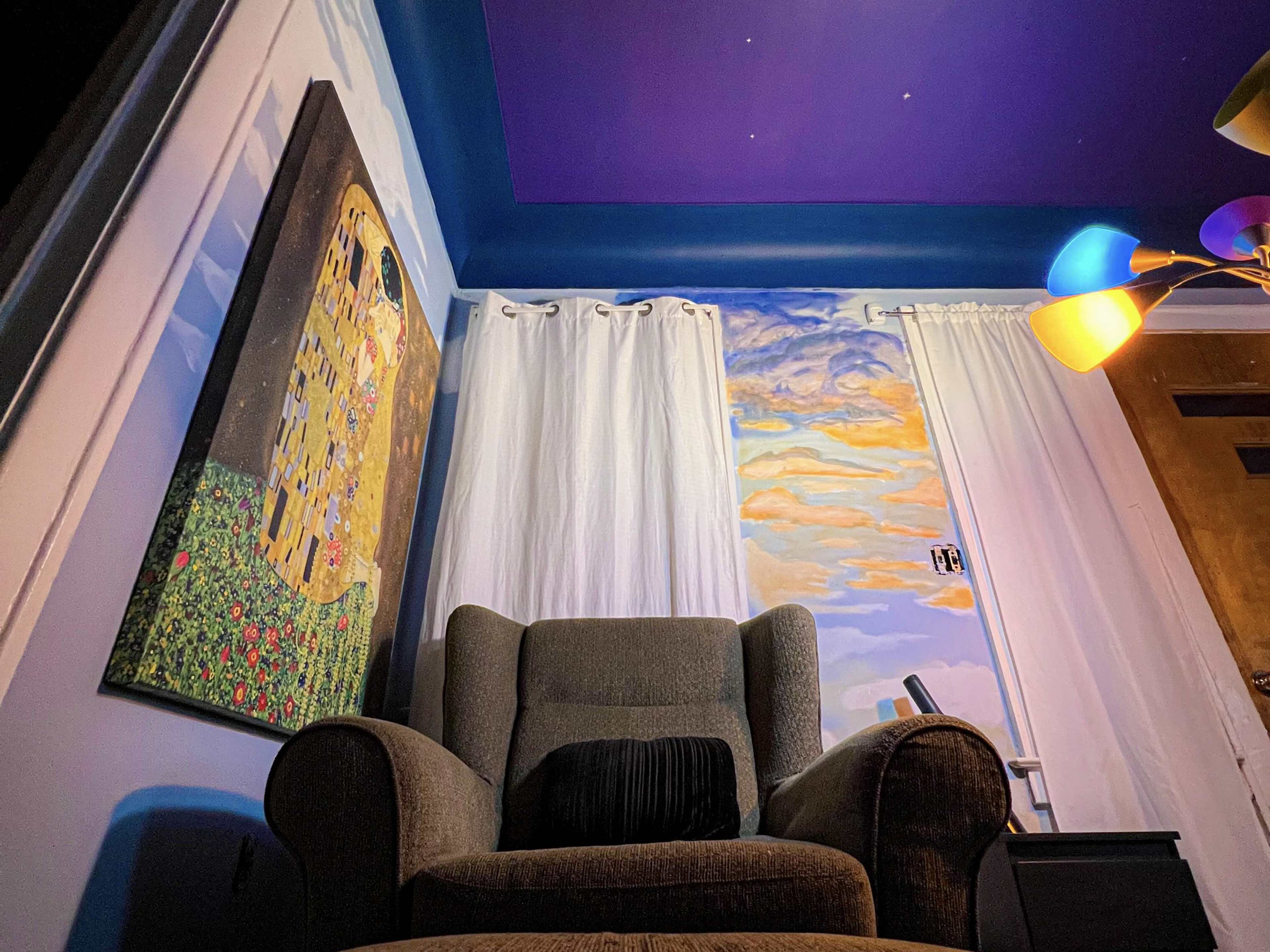 A cozy corner of a room features a green armchair, colorful lamp, and walls adorned with a large painting and bright curtains.