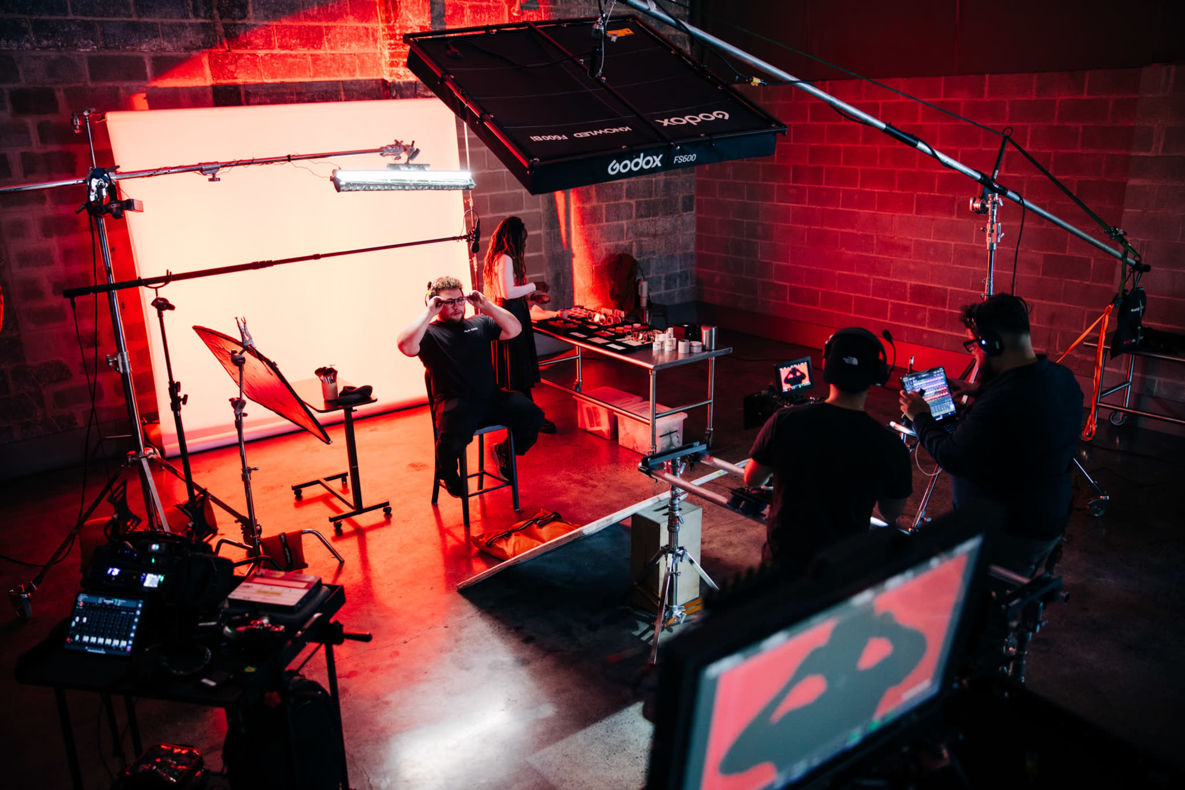A studio setup features a person sitting on a stool in front of a lit backdrop, with cameras and lighting equipment arranged around them.