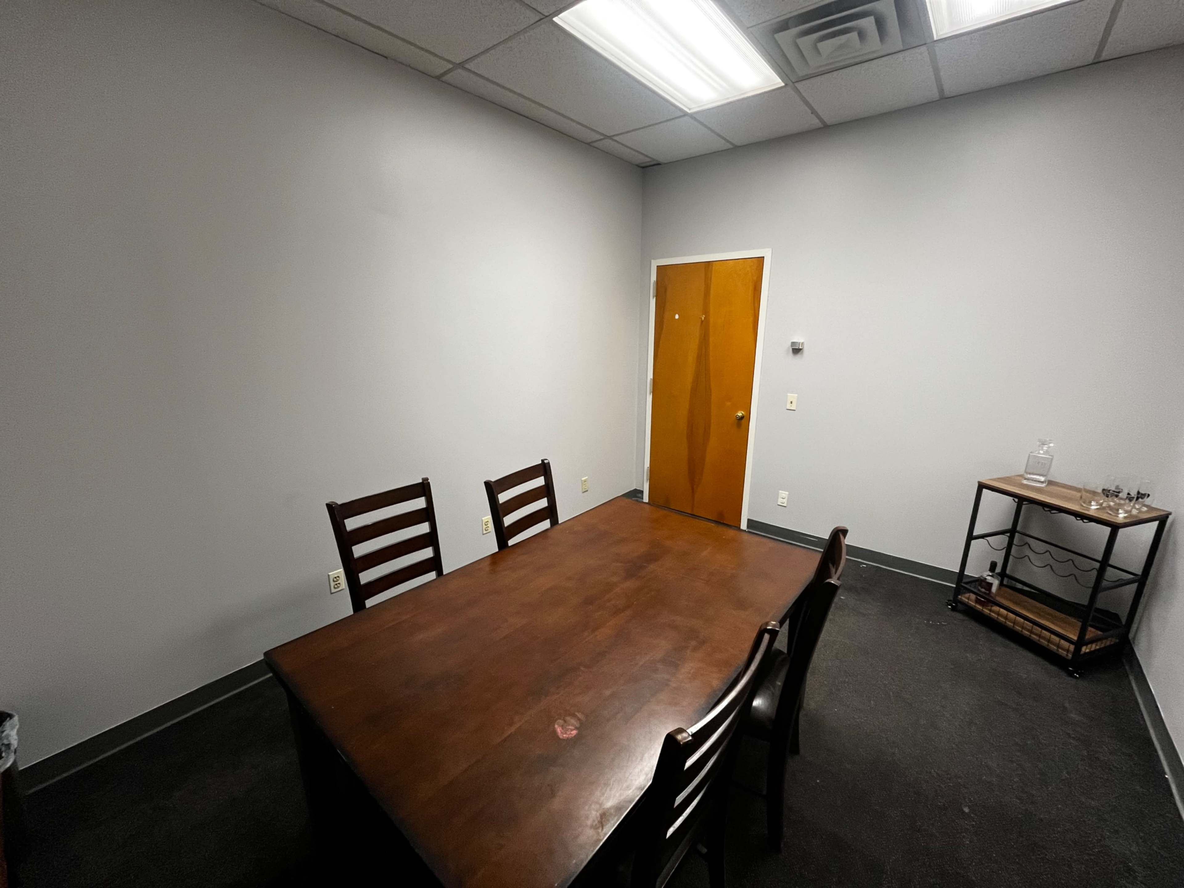 An empty conference room features a large wooden table with four chairs and a side cart with glassware.