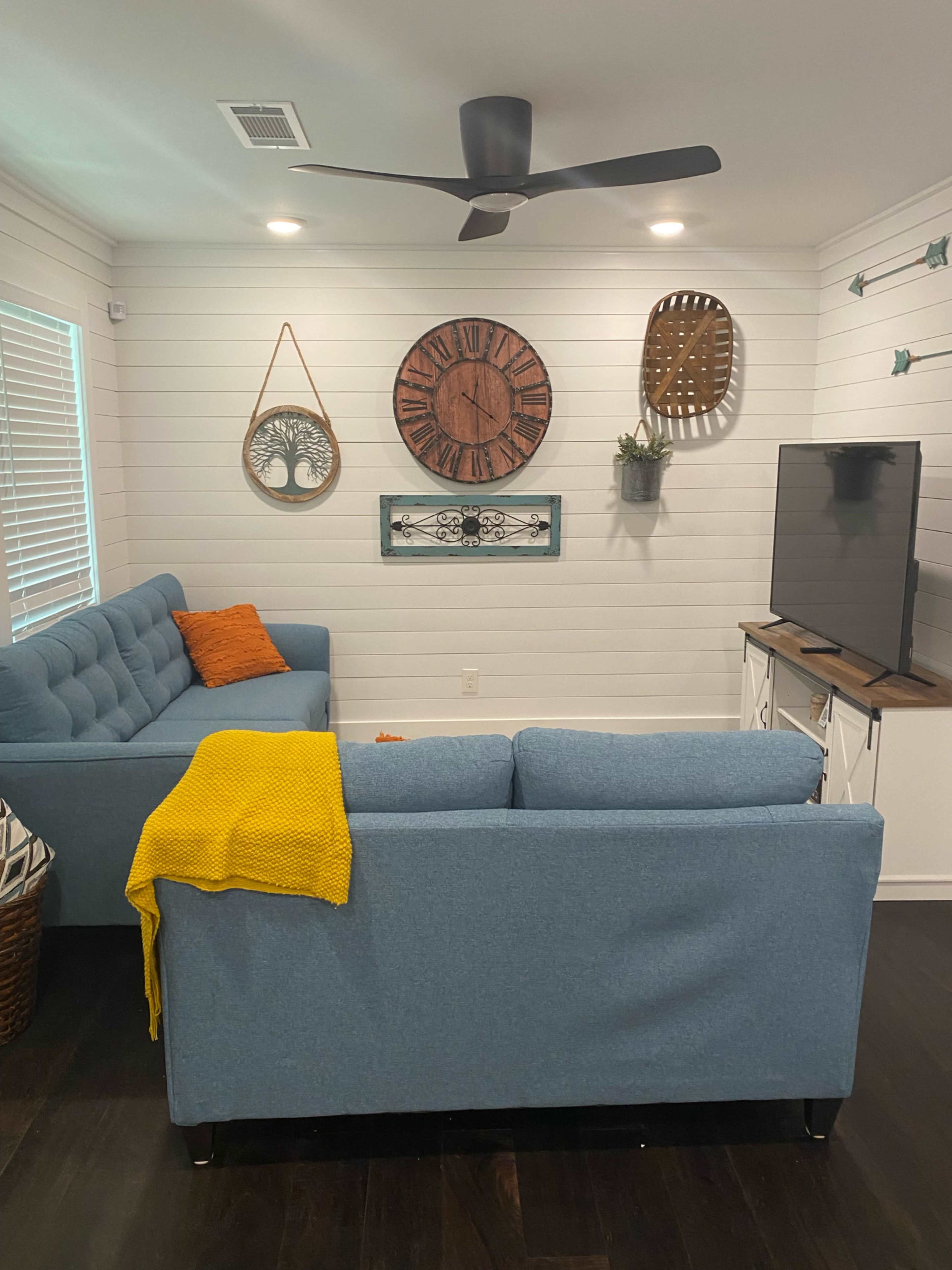 A modern living room features a blue sectional sofa, a wooden wall clock, decorative wall hangings, and a flat-screen TV.