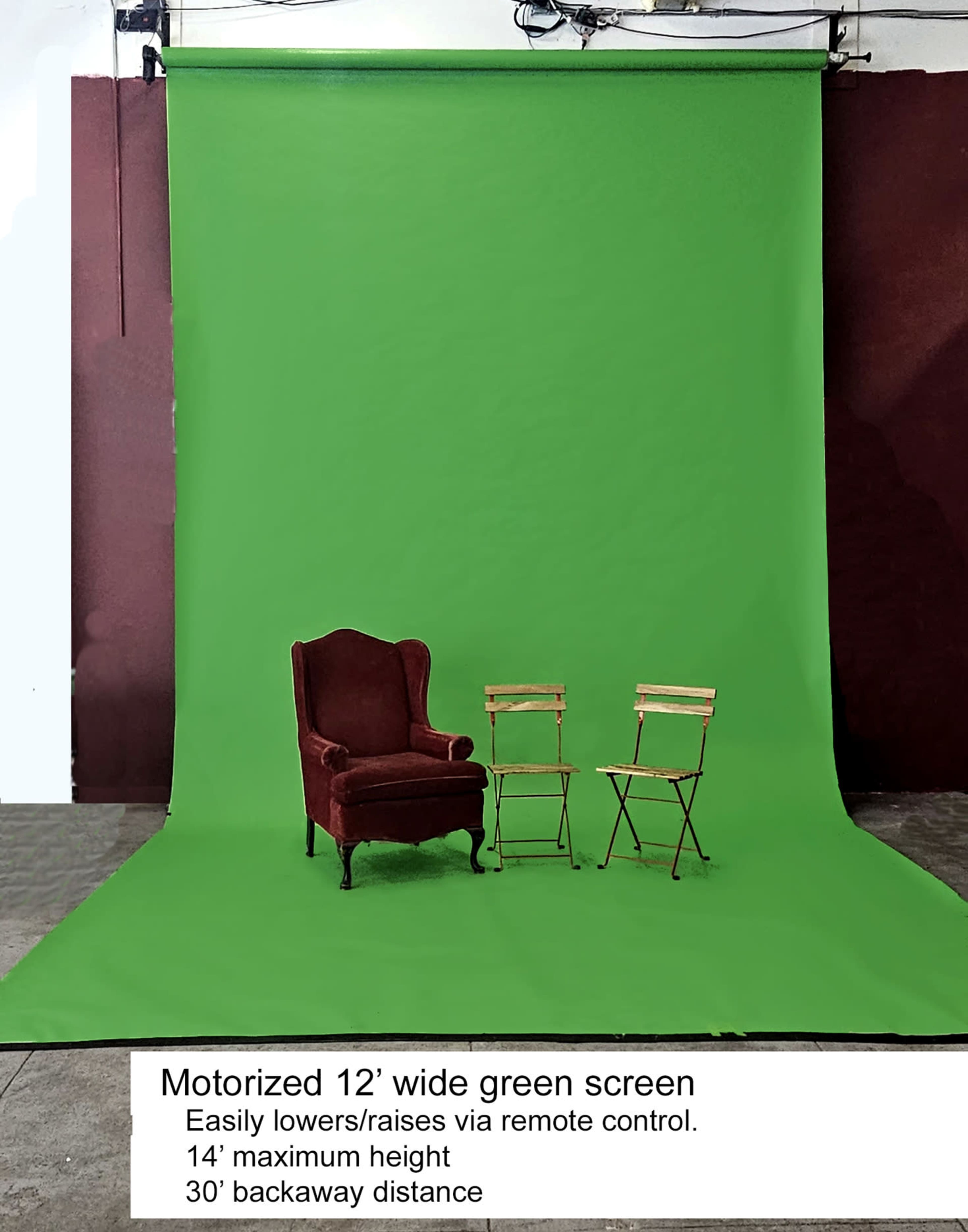 A motorized 12-foot wide green screen set up in an indoor space, accompanied by a red upholstered armchair and two wooden director's chairs.