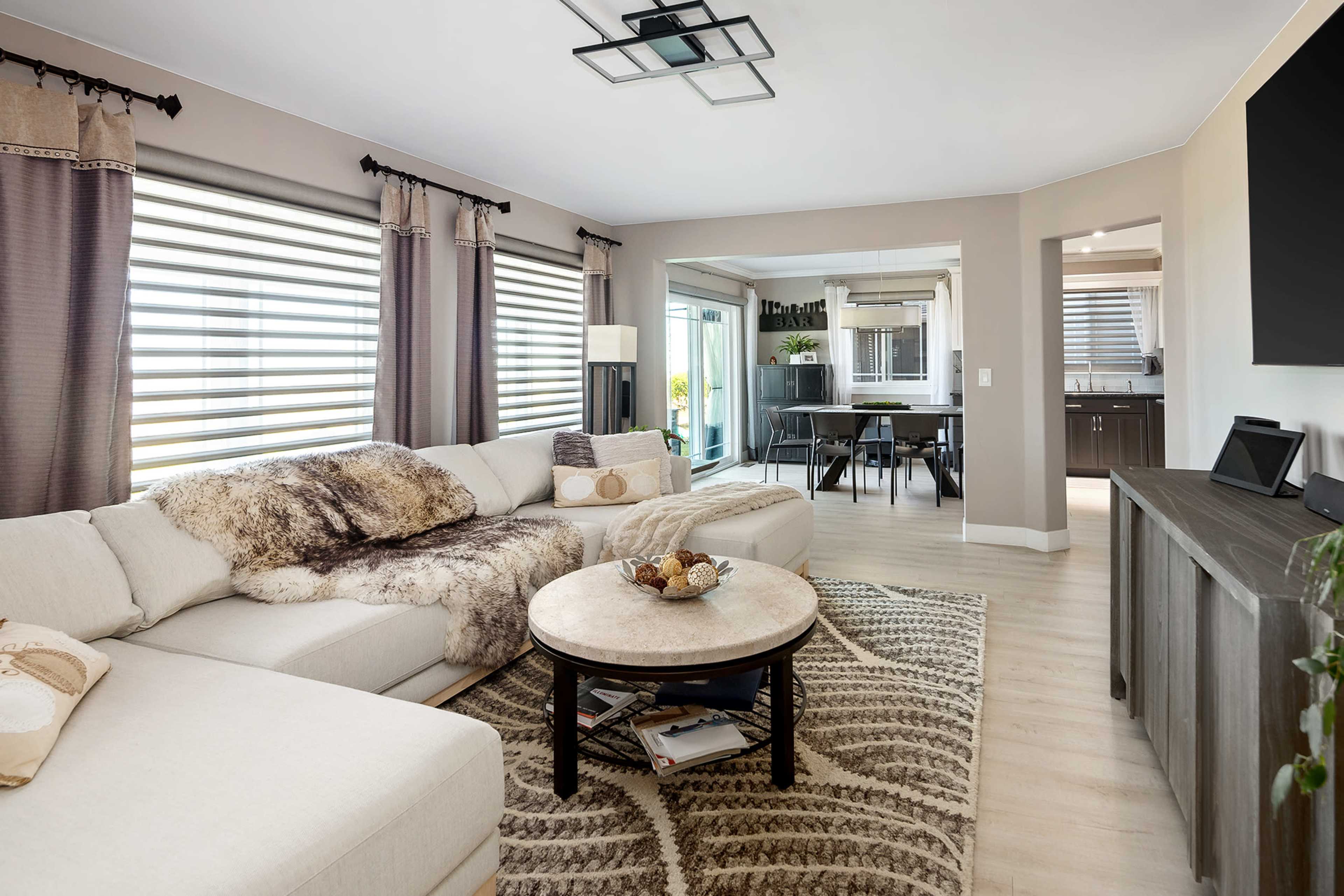 A modern living room features a large sectional sofa, a round coffee table, and large windows with blinds, connecting to a dining area visible in the background.