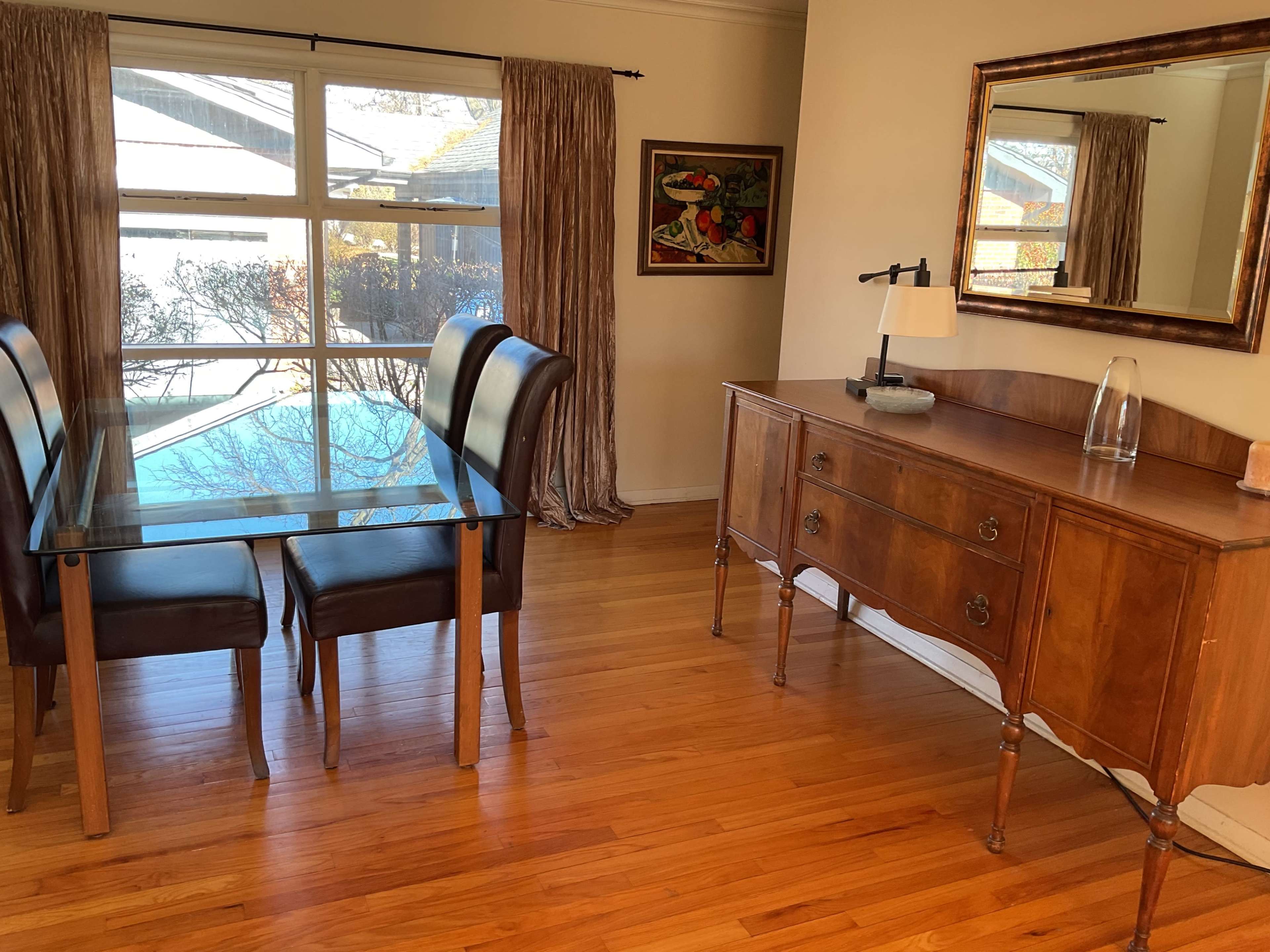 The image shows a dining area featuring a glass-top table with two chairs and a wooden sideboard, complemented by a large window with curtains and a framed painting on the wall.