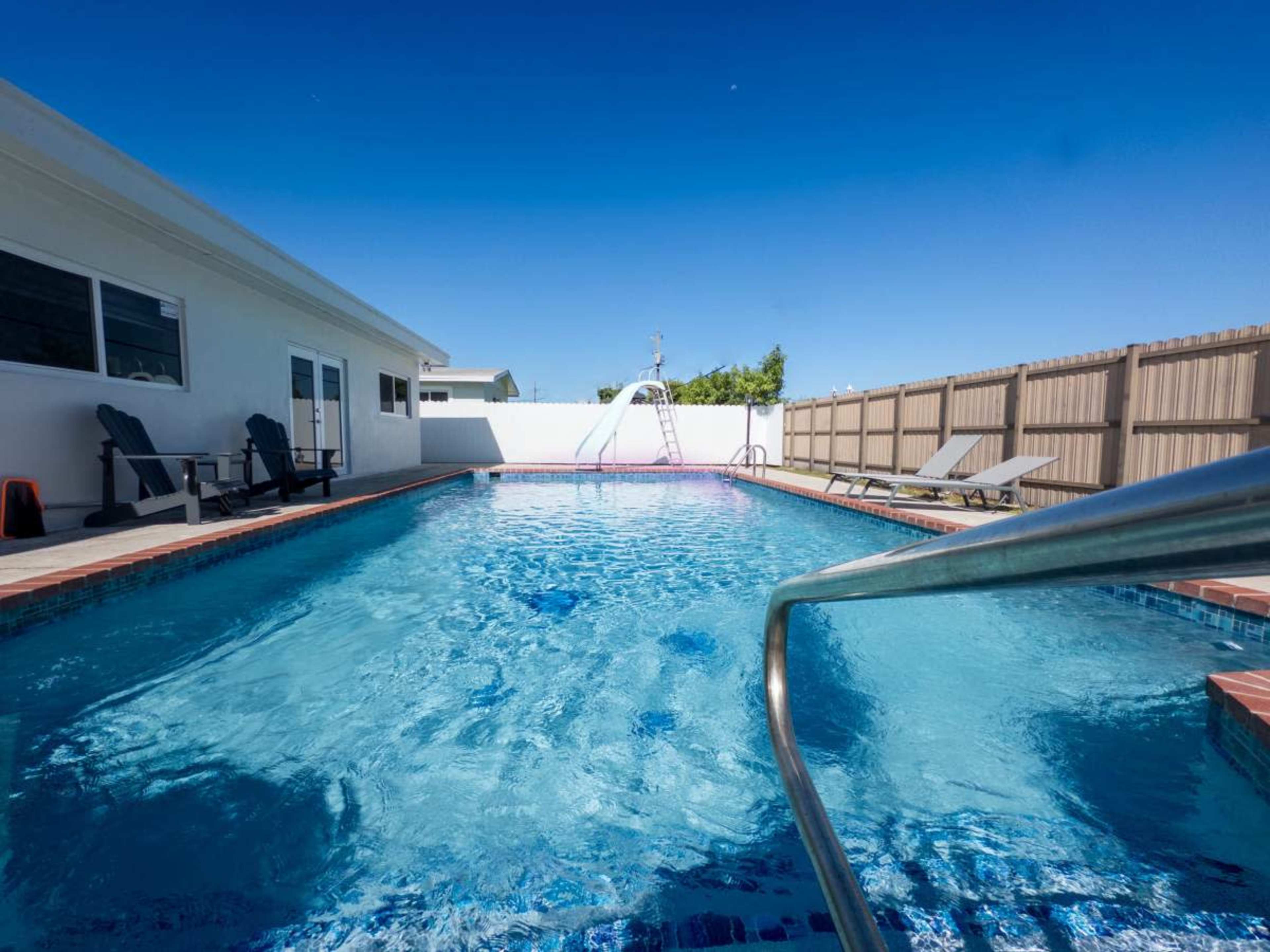A clean, rectangular swimming pool is surrounded by lounge chairs and a wooden fence under a clear blue sky.