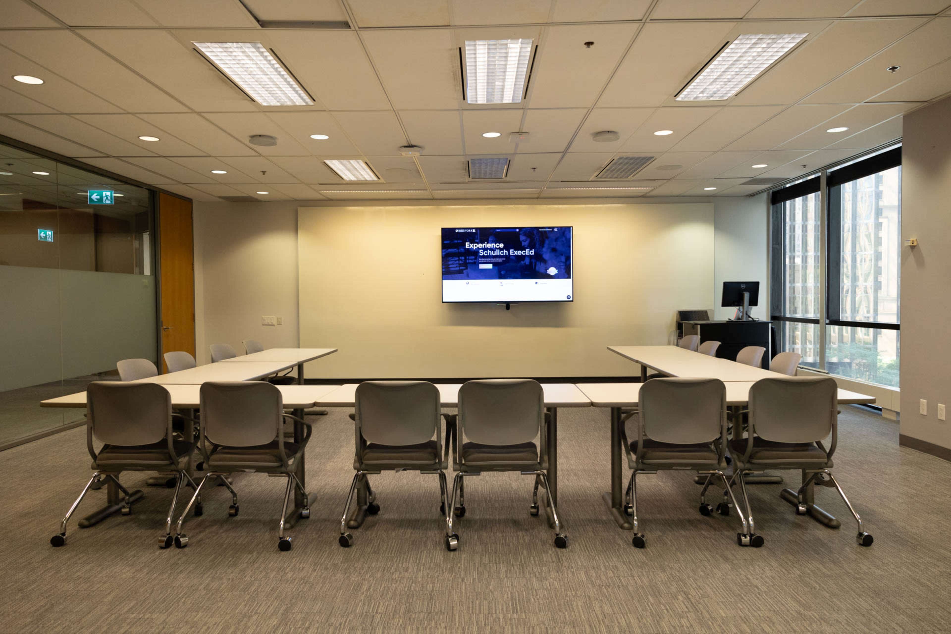 A modern conference room features a central table surrounded by chairs, with a large screen mounted on the wall.