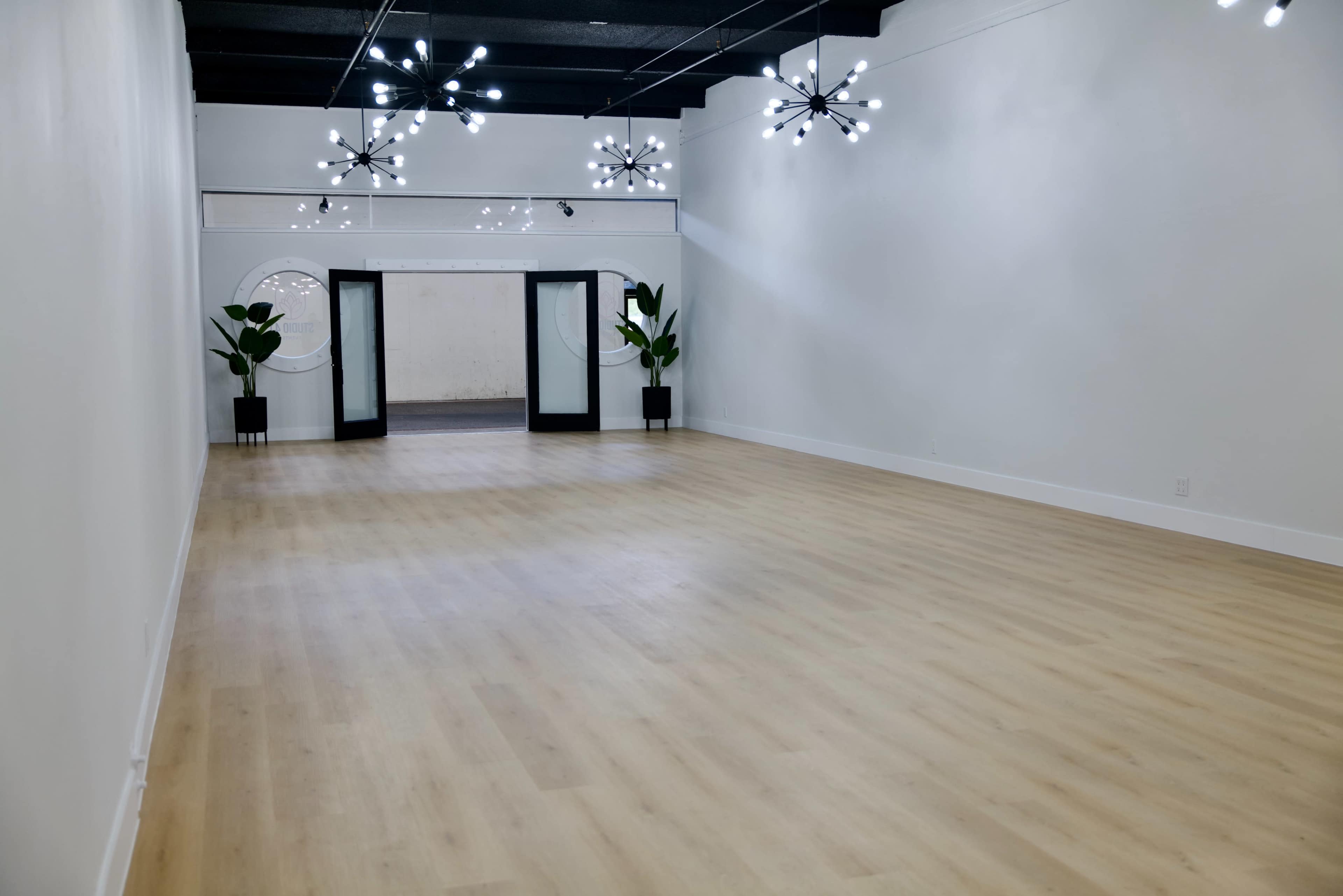 The image shows a spacious, empty room with light wooden flooring, featuring modern chandeliers and two large glass doors leading to another area.