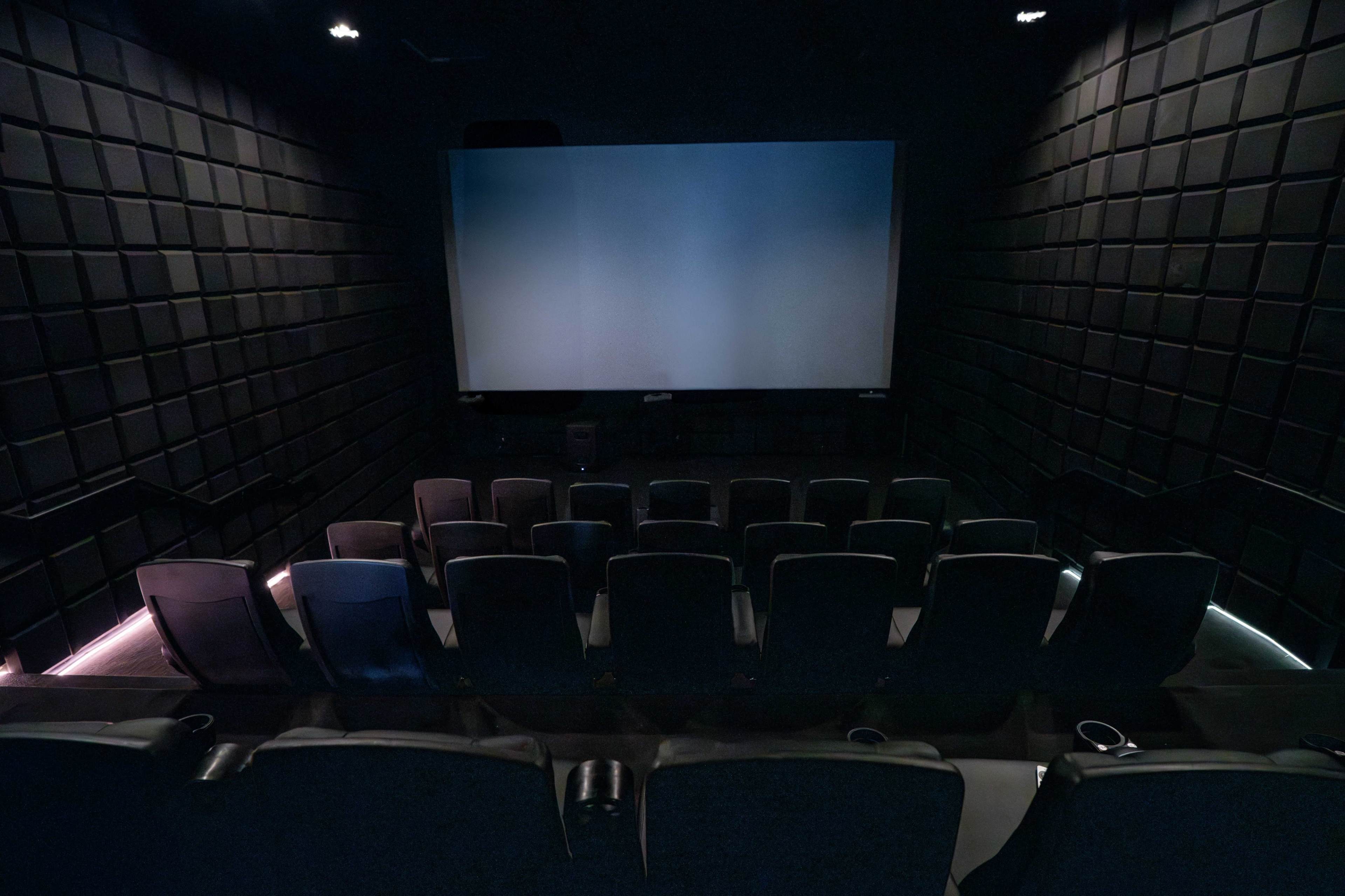 The image shows a darkened cinema with empty rows of black seats facing a large, blank screen.